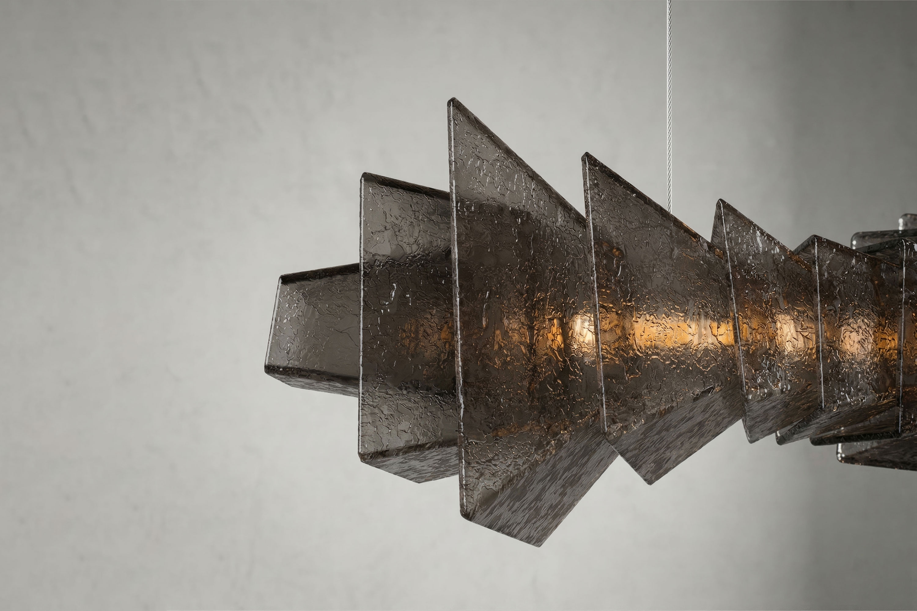Close-up of a modern geometric pendant light, handmade with textured, smoked Murano glass panels arranged in a zigzag pattern, hanging against a plain, light gray wall. A warm light glows from the center.