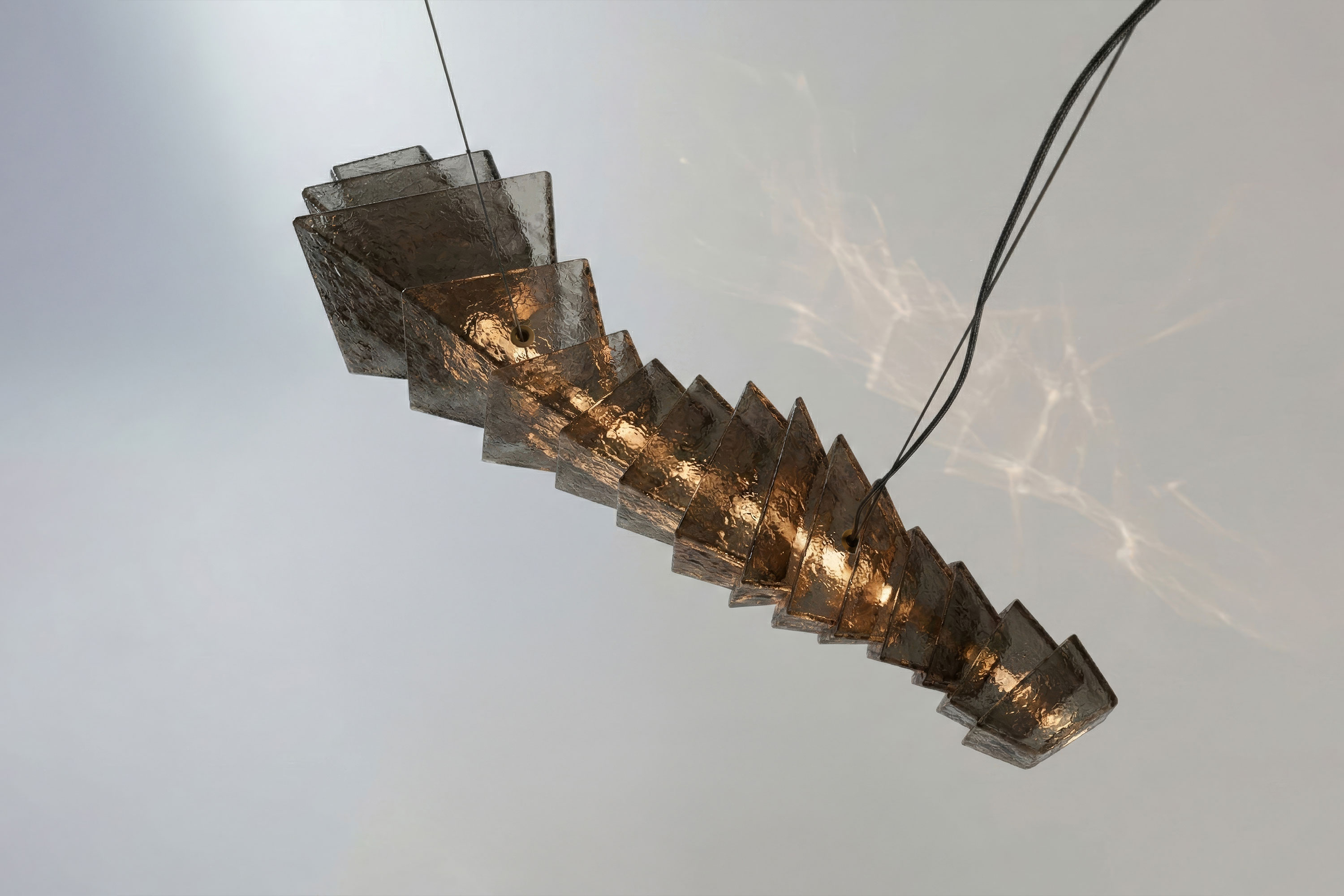 A sculptural hanging light fixture, handmade from stacked, angled translucent panels with embedded bulbs, is suspended by two cables against a plain background.