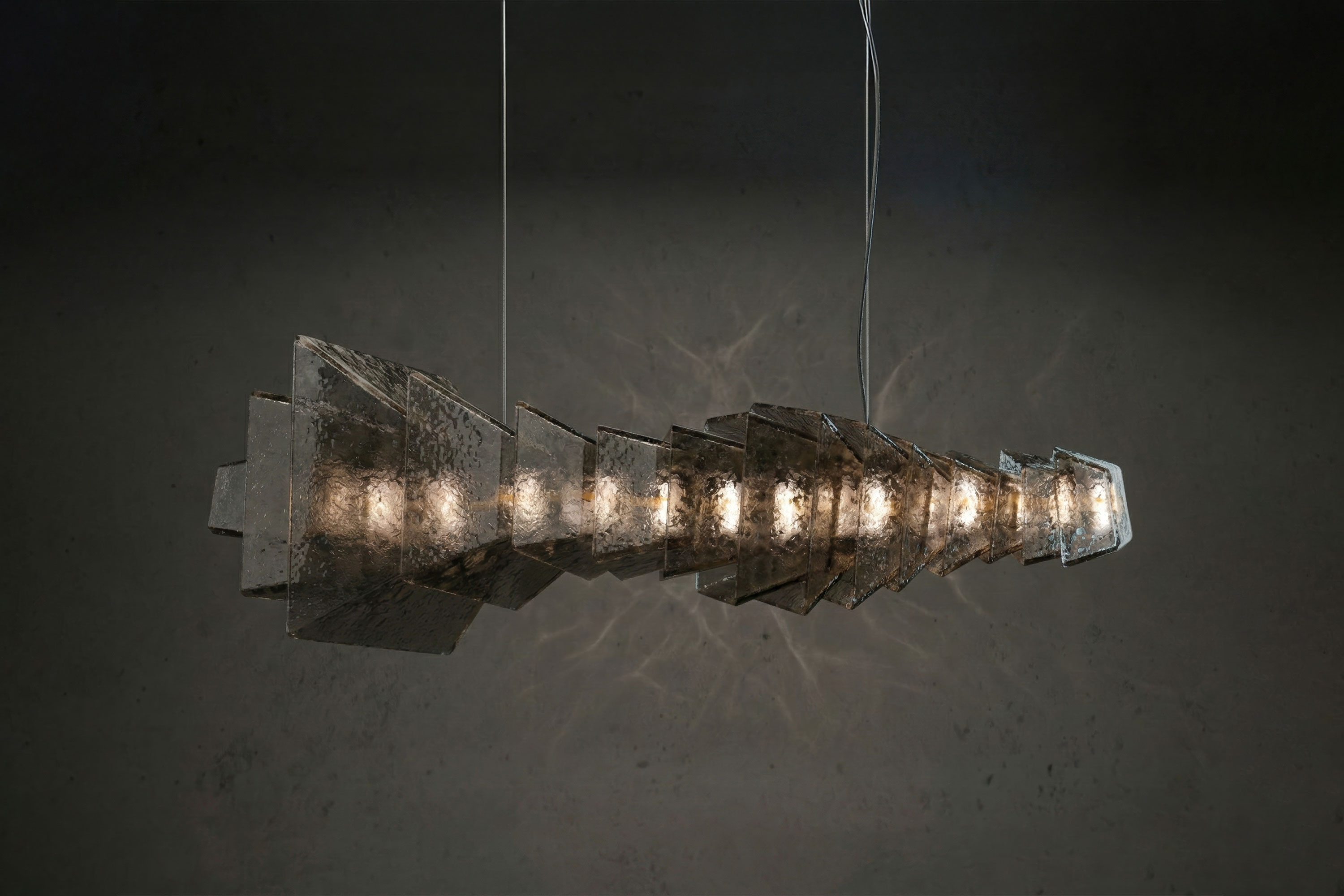 A modern chandelier with multiple rectangular, metallic panels arranged in a spiral shape. Visible light bulbs are evenly spaced along the center. The fixture, reflecting the Art Deco style, hangs from thin wires against a dark gray background.