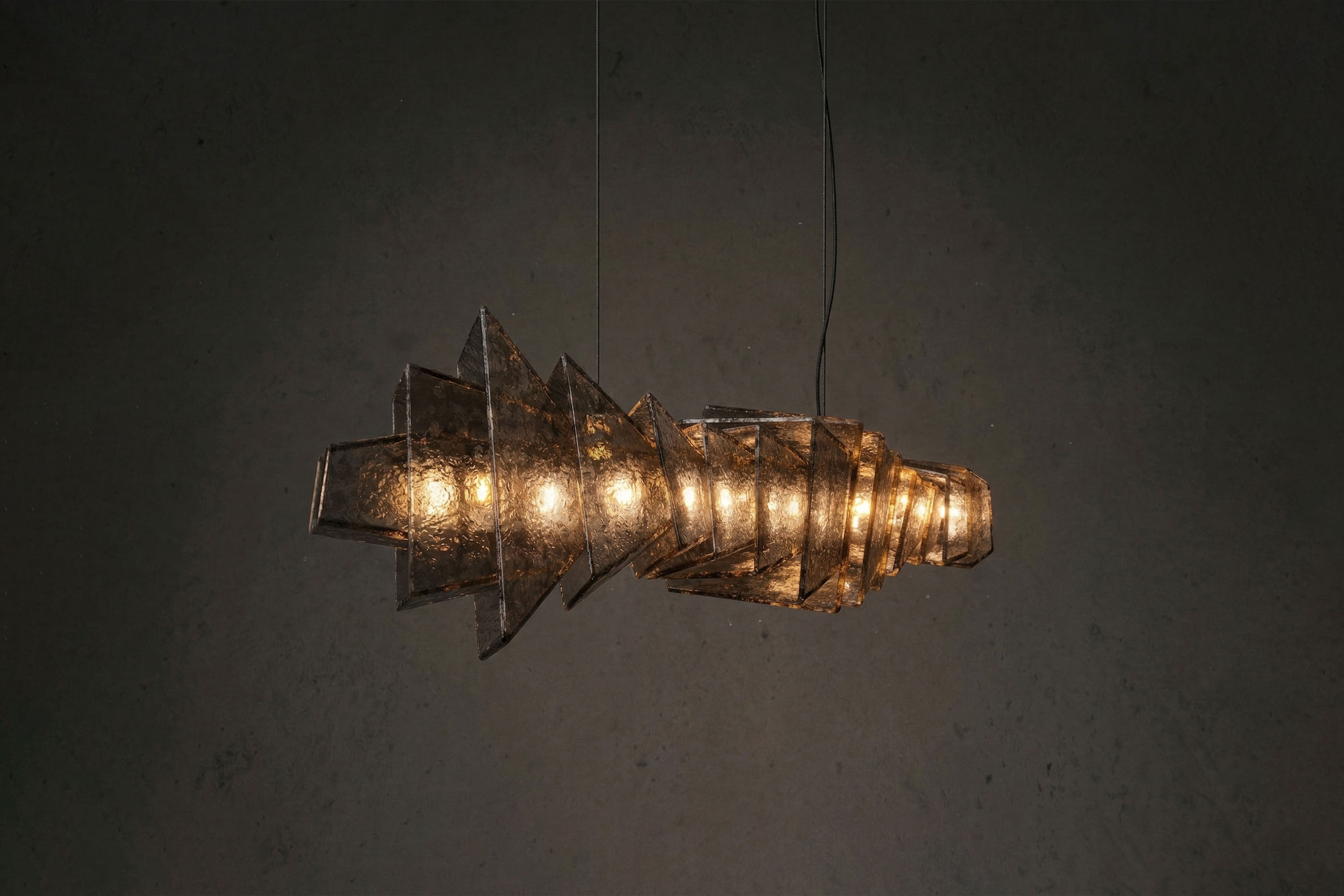 A geometric pendant light fixture with angular, overlapping brown panels emits a warm glow against a dark background. Handmade craftsmanship is evident in the design, and two cables suspend the fixture from above.