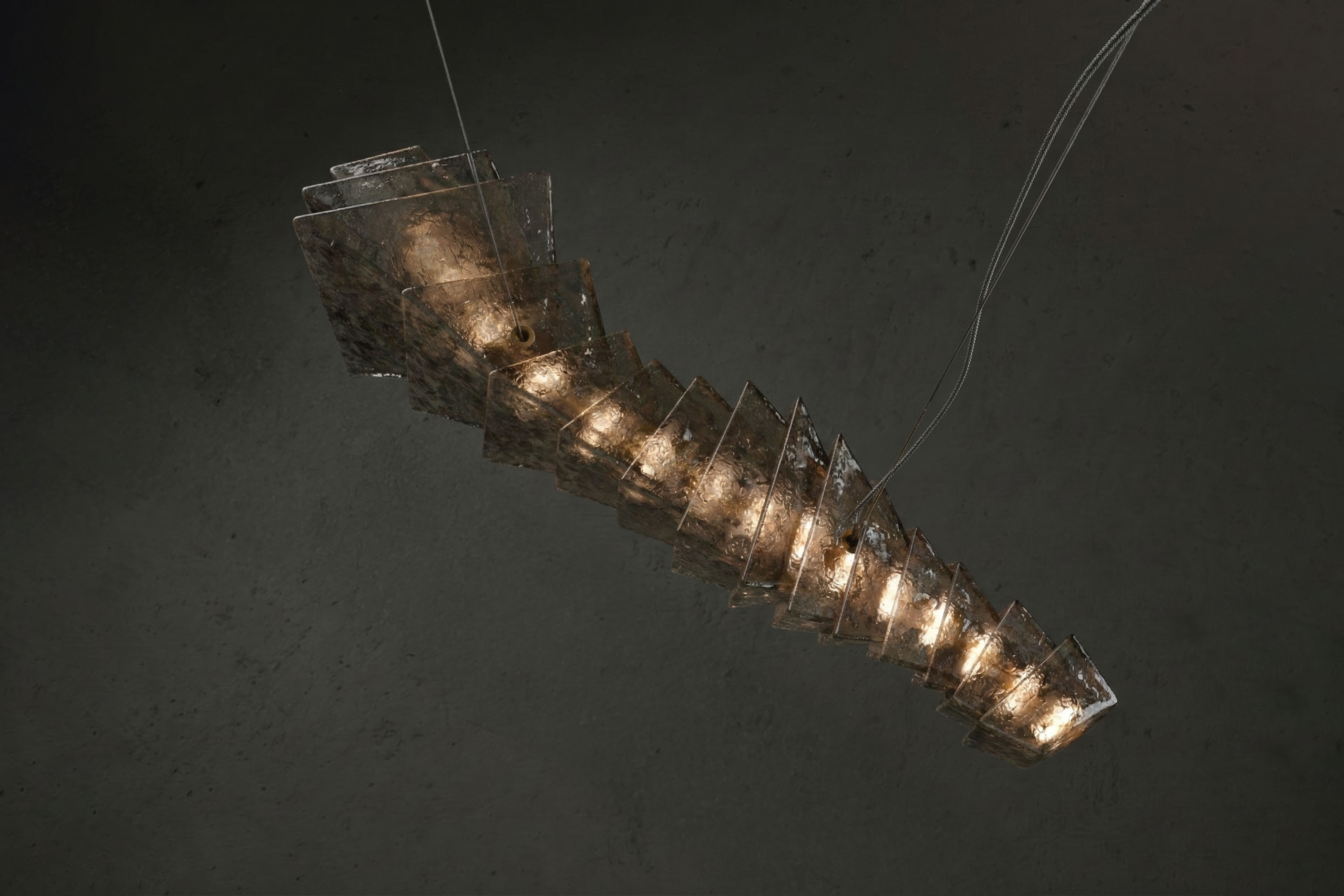 A modern chandelier featuring layered, tilted rectangular Murano glass panels surrounds internal lights, hanging at an angle against a dark background; two suspension wires are visible.
