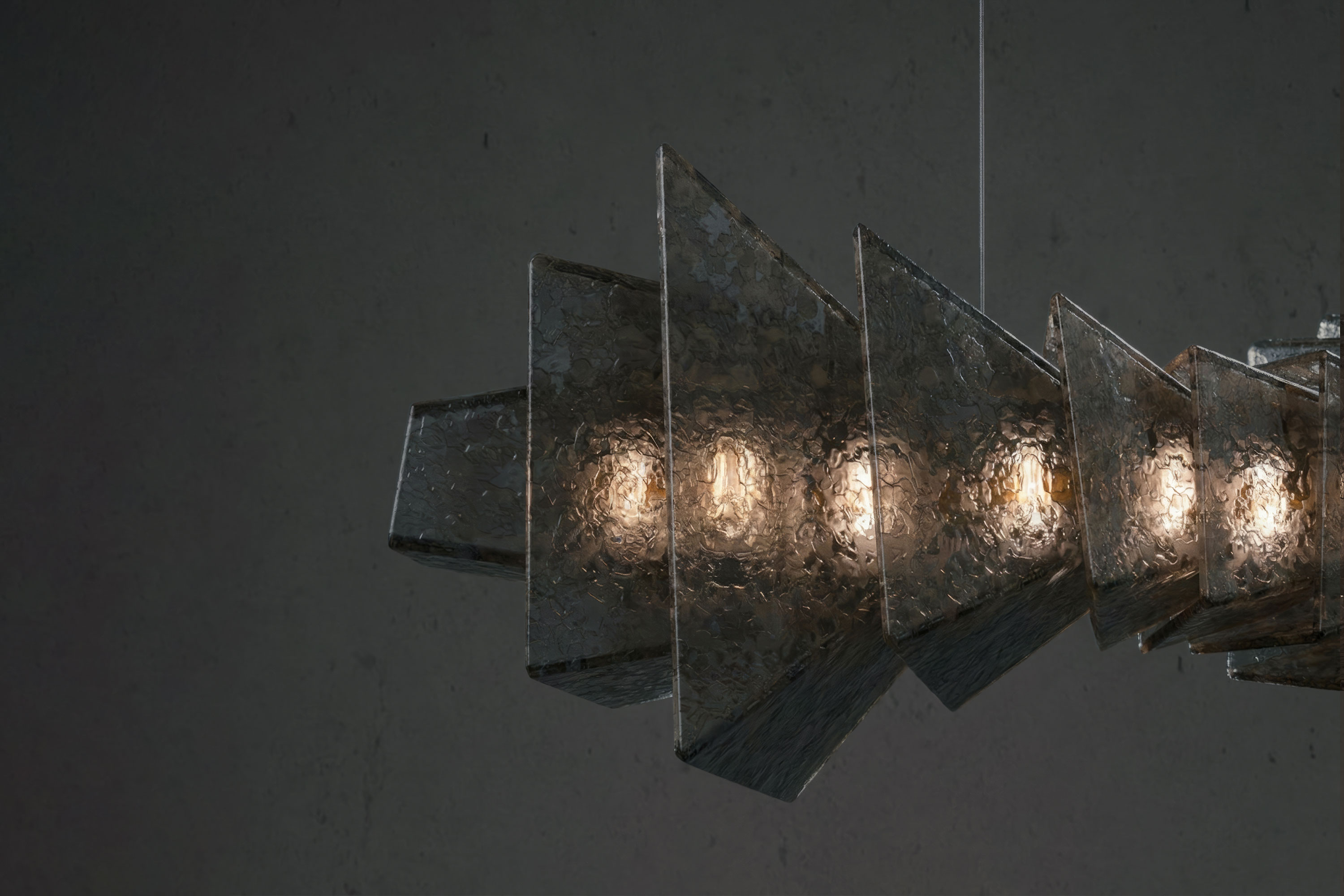 A modern bronze metal pendant light with angular, overlapping panels and exposed bulbs, suspended from the ceiling against a dark, plain background.