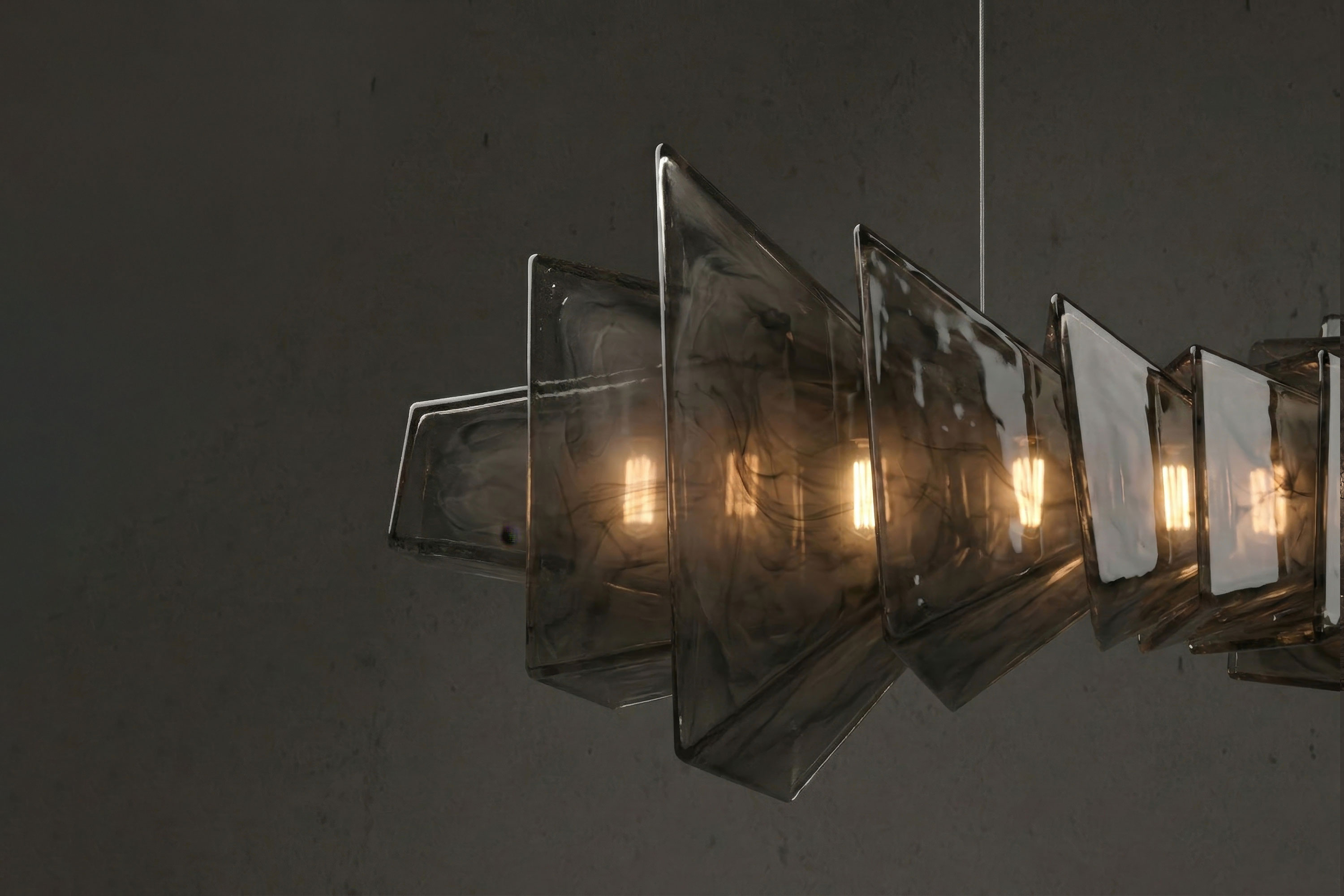 Modern pendant light fixture with multiple handmade glass panels arranged in a geometric, overlapping pattern. Warm lightbulbs are visible inside, casting a soft glow. The background is a plain, dark grey wall.