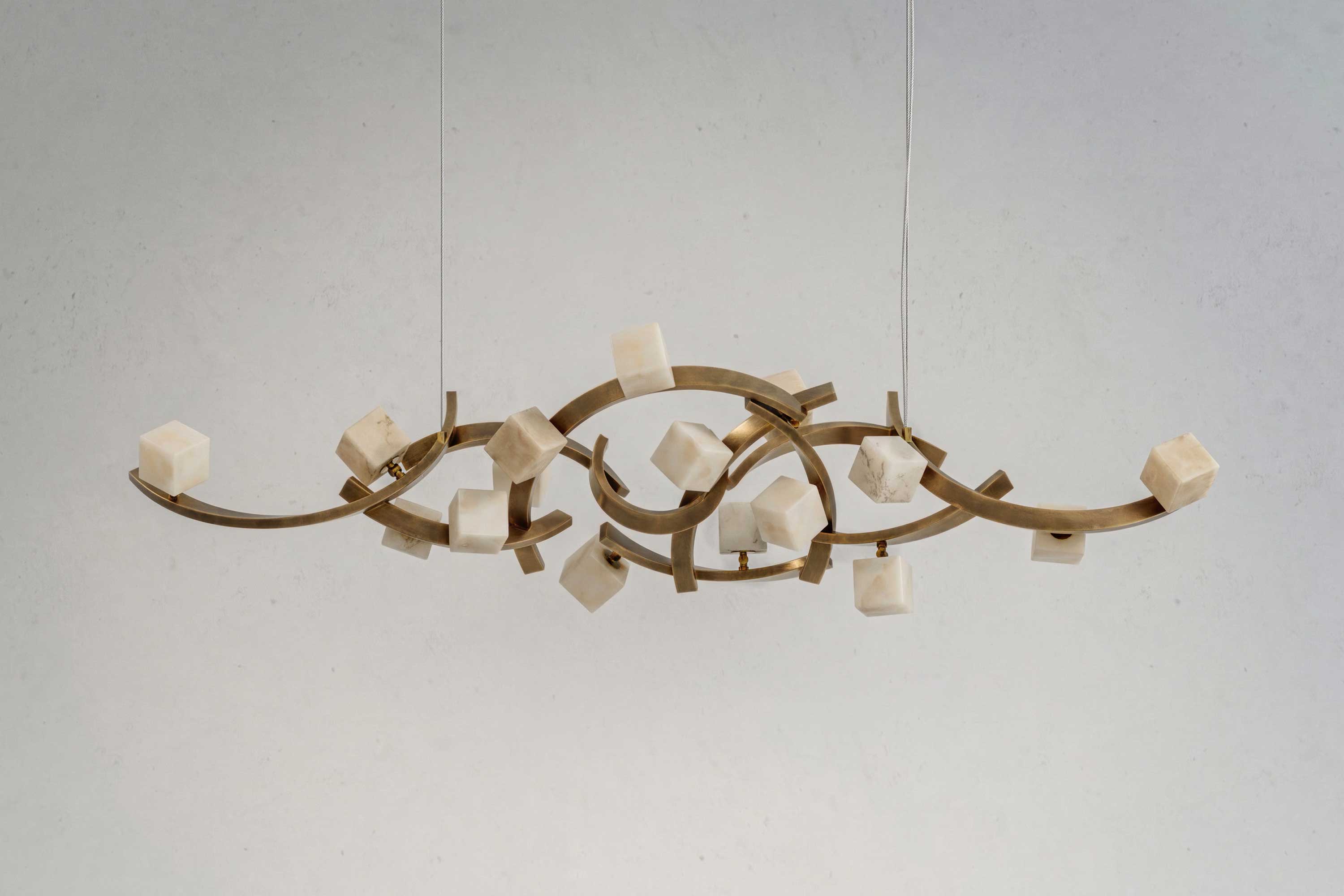 A modern Artdecoo chandelier with a curving bronze frame features several white stone-like cubes attached at different points, suspended by two thin wires against a plain light gray wall.