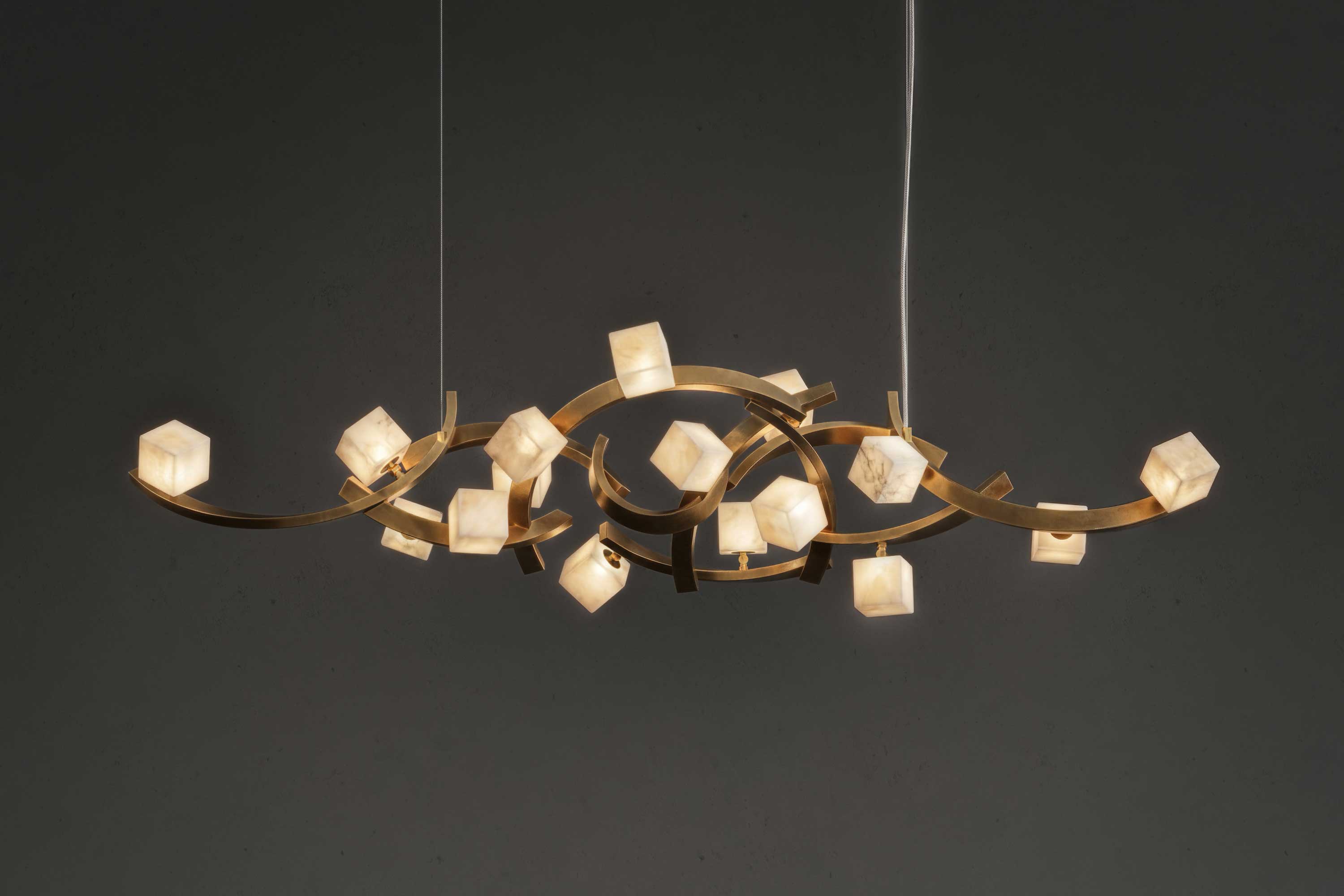 A modern chandelier with a gold-colored metal frame and multiple glowing cube-shaped lights, suspended by two cords against a dark background. This piece features handmade detailing reminiscent of bespoke furniture, enhancing its unique appeal.