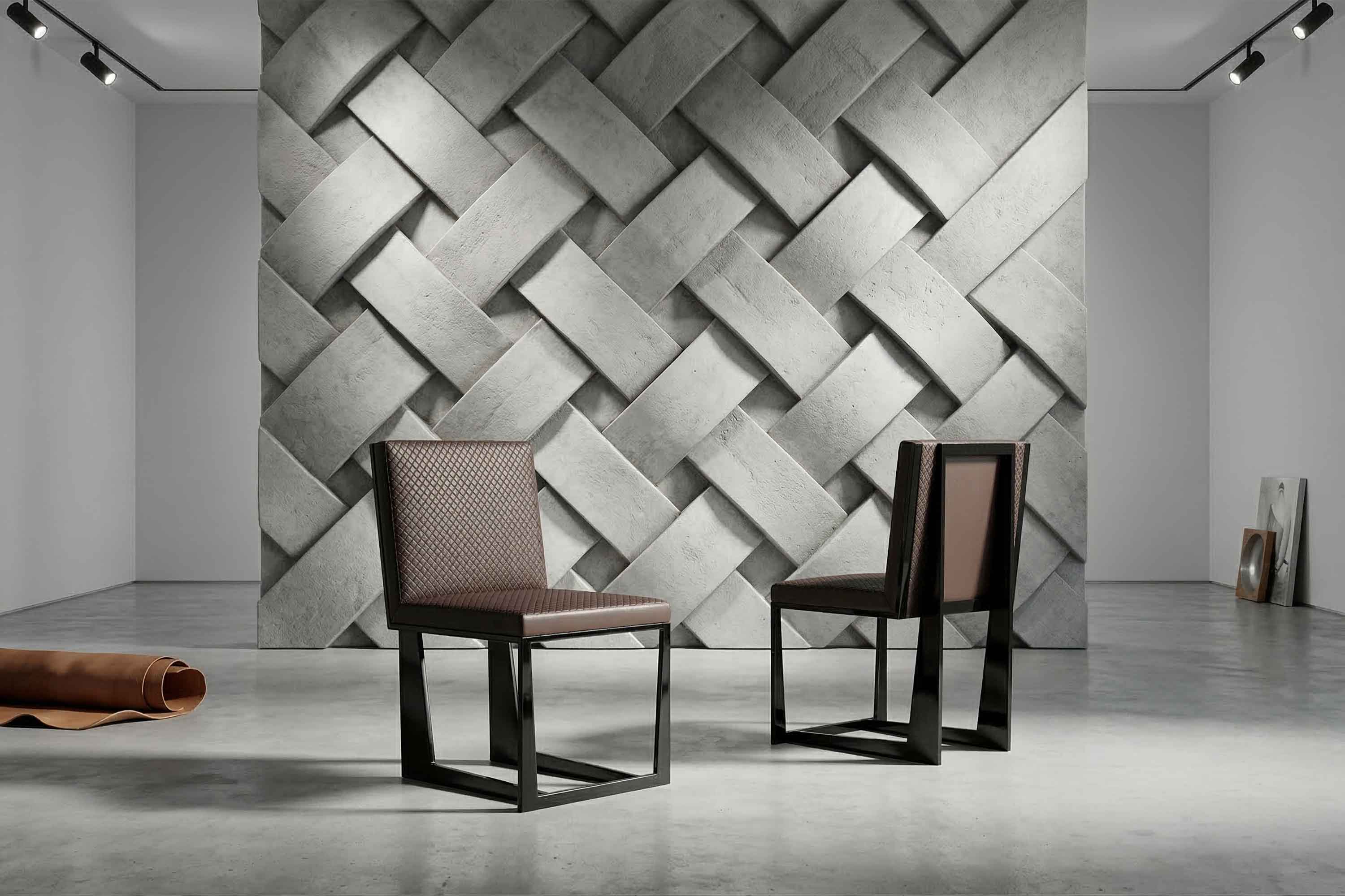Two modern chairs with metal frames are positioned on a polished concrete floor in front of a large, textured, woven-pattern gray wall. Rolled-up material and framed artwork rest against the walls in the minimalist room.