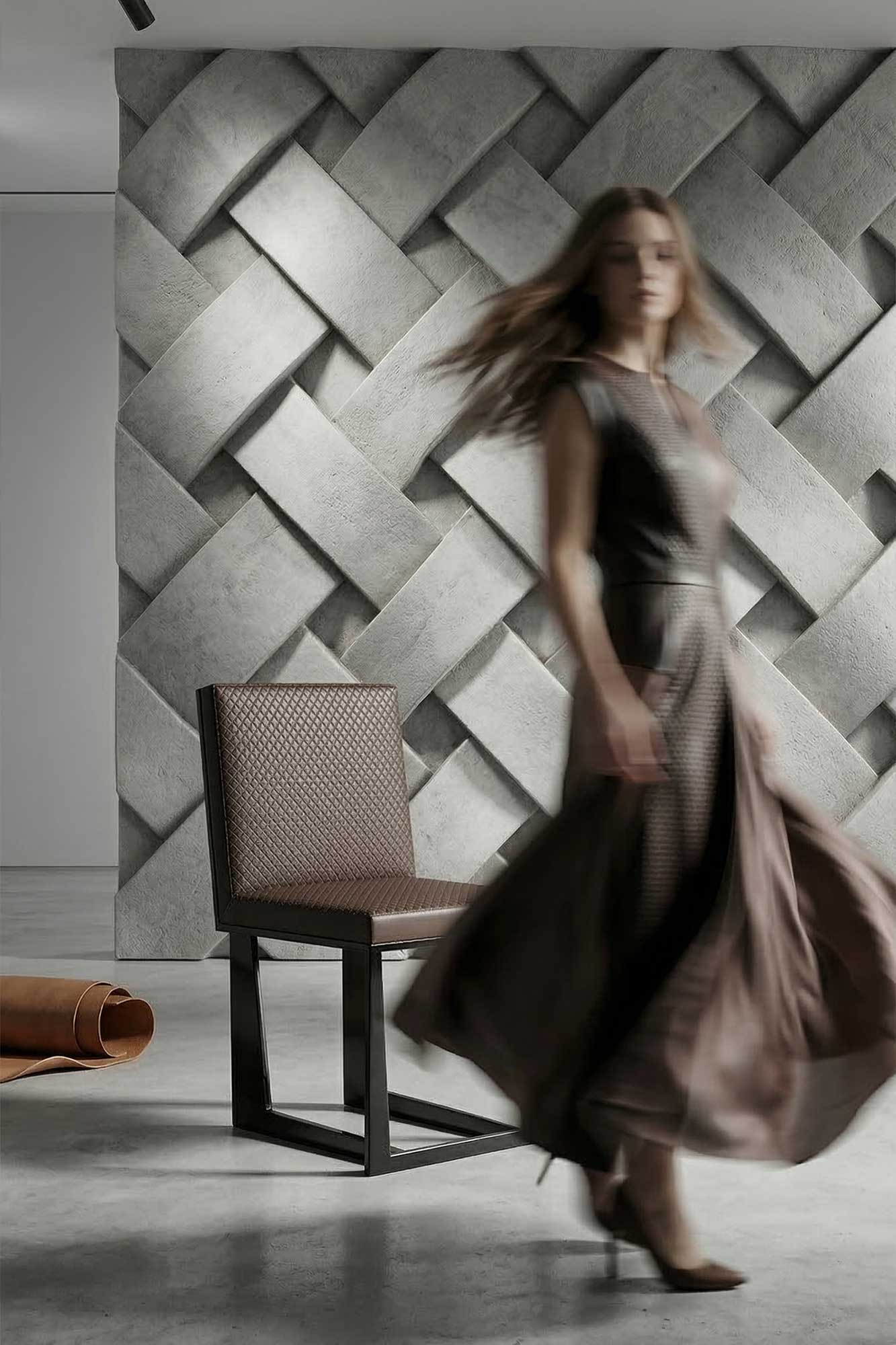 A woman in a flowing dress walks past a chair in front of a textured, woven-pattern wall. A rolled-up material lies on the floor nearby. The woman appears blurred, suggesting motion.