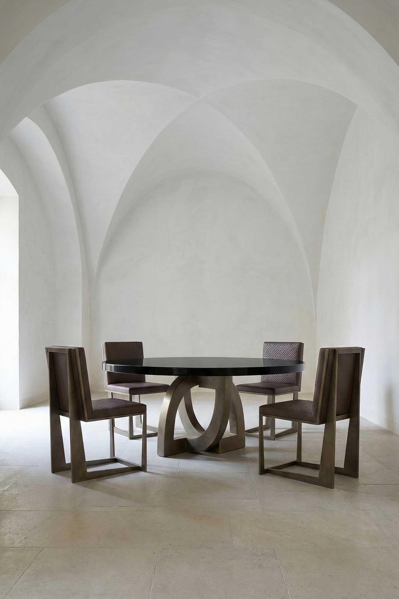 A round black table with a sculptural base is surrounded by four angular wooden chairs in a minimalist room with vaulted white ceilings and pale stone floors.