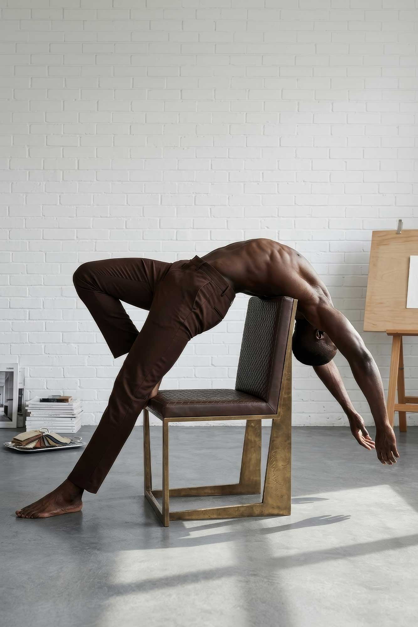 A shirtless person in brown pants arches their back dramatically over a square chair in a bright room with white brick walls, books, and an easel in the background.