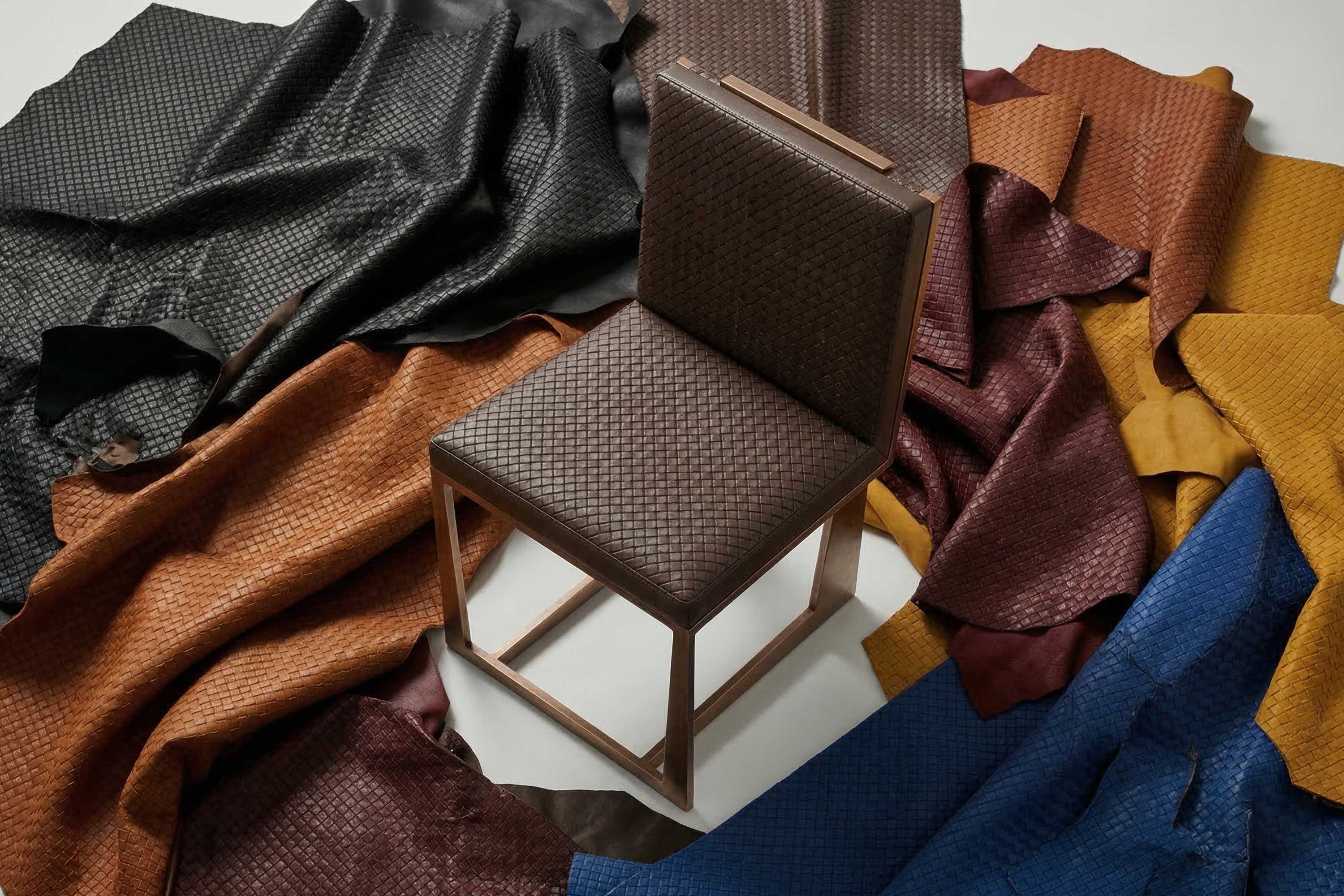 A modern wooden chair with dark brown woven upholstery is placed on a white surface, surrounded by large sheets of textured leather in black, brown, red, mustard yellow, and blue.