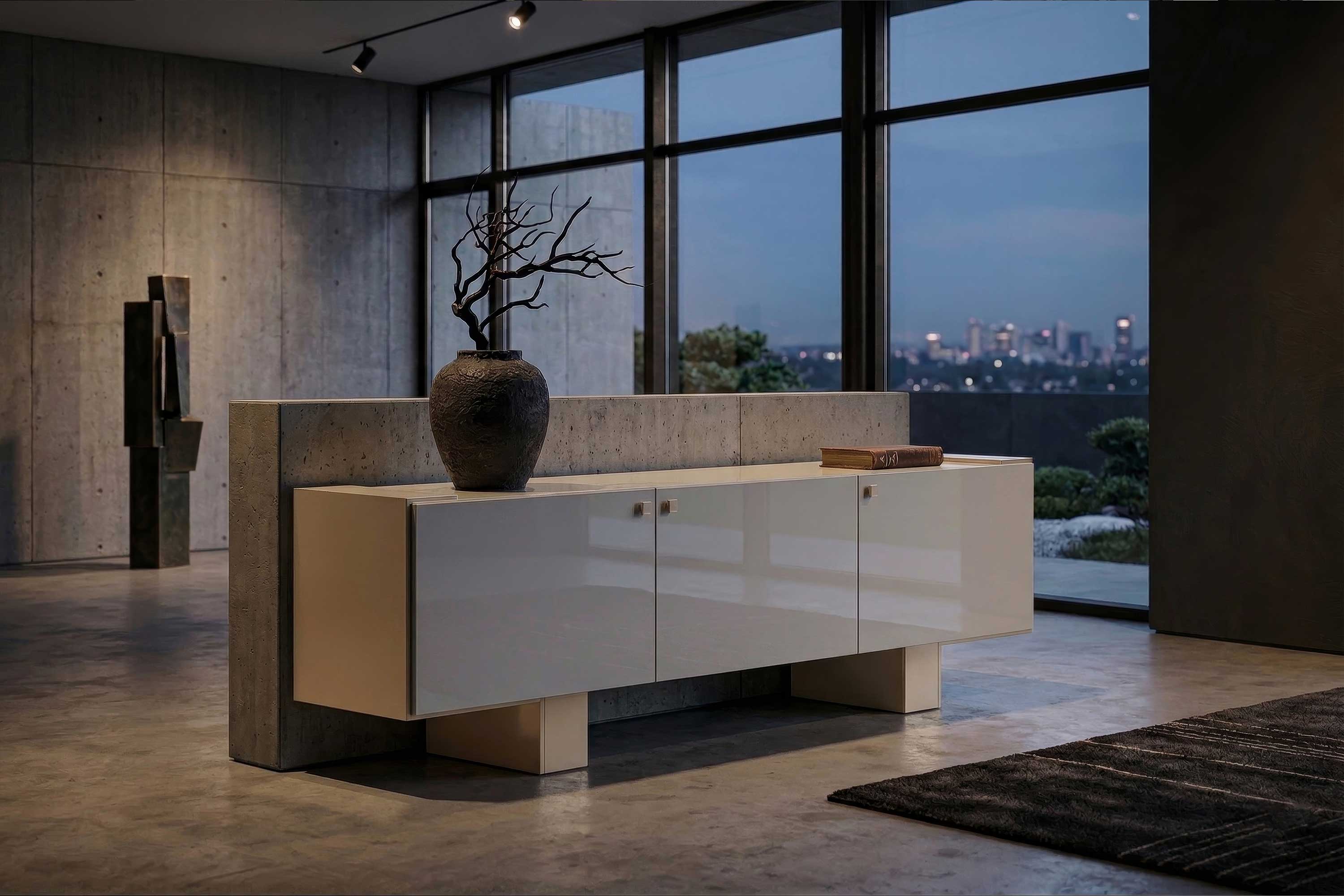A modern room with a glossy white sideboard, a large vase with bare branches, a book, concrete walls, large windows, and a cityscape visible outside at dusk.