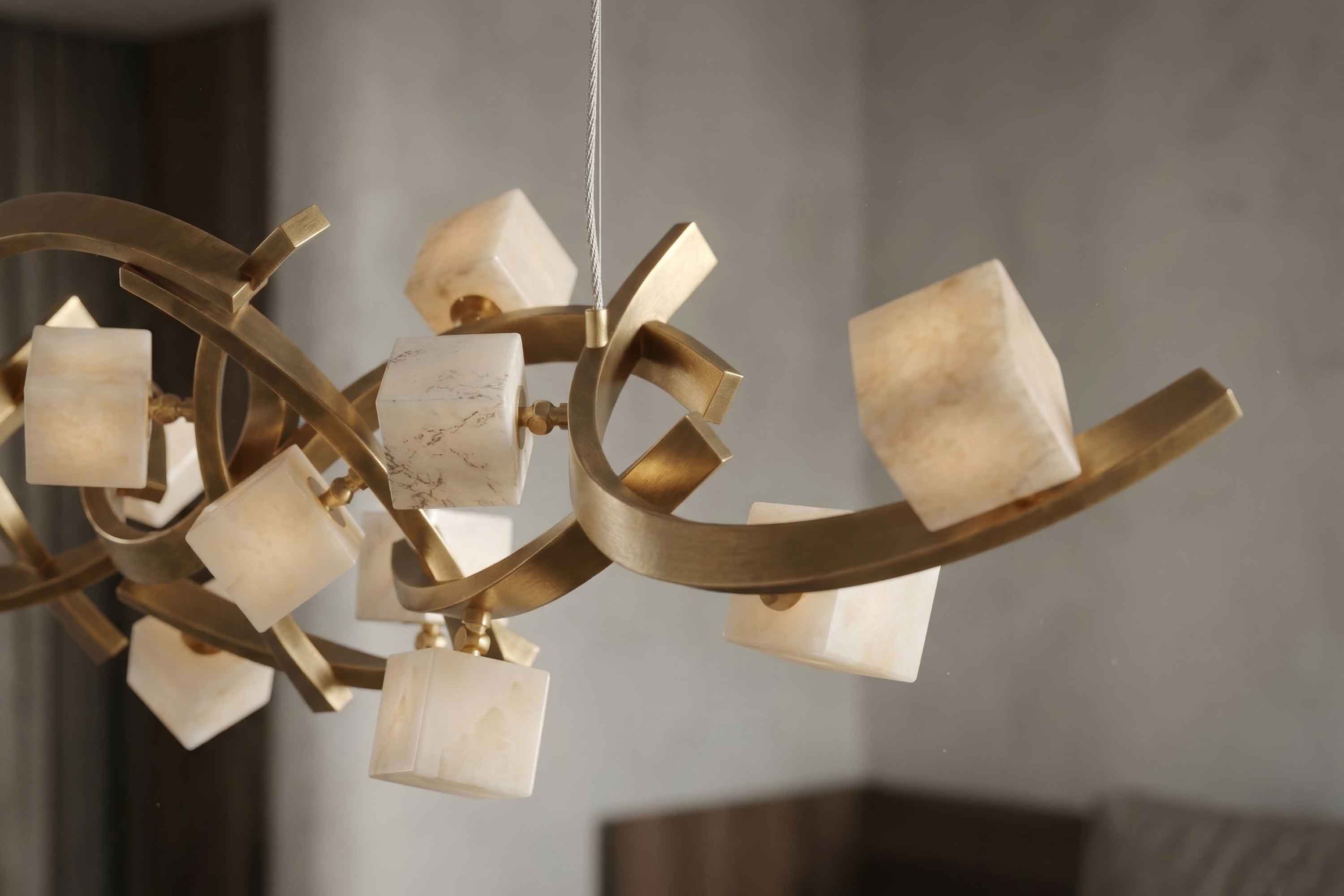 A modern chandelier features gold-colored metal curves with multiple cube-shaped white stone or frosted glass shades, mounted on a ceiling with a neutral, blurred background.