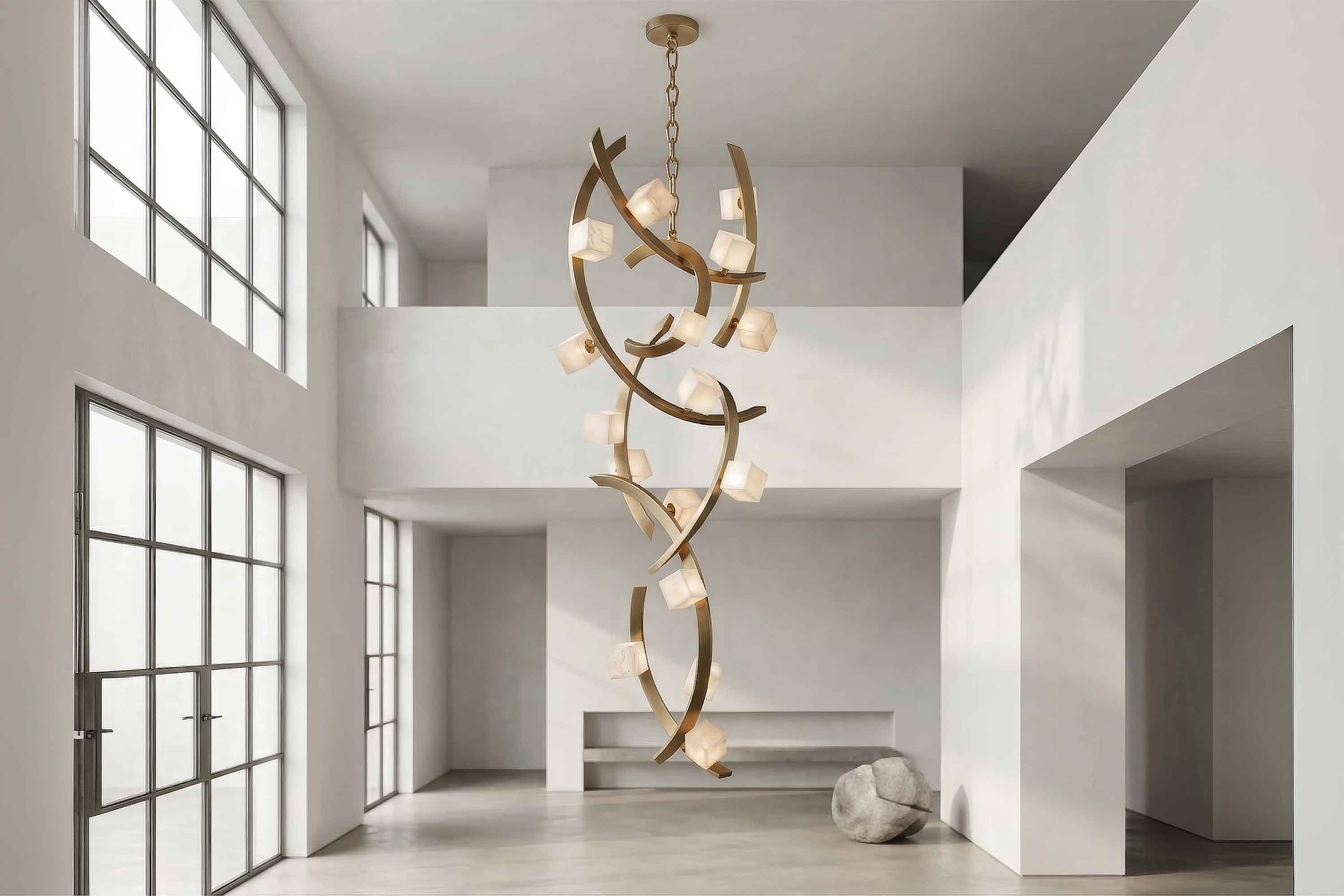 Large, modern chandelier with gold, branch-like structure and cube-shaped lights hangs in a spacious, minimalist room with white walls, high ceilings, large windows, and polished concrete floors. A round stone sits in the corner.