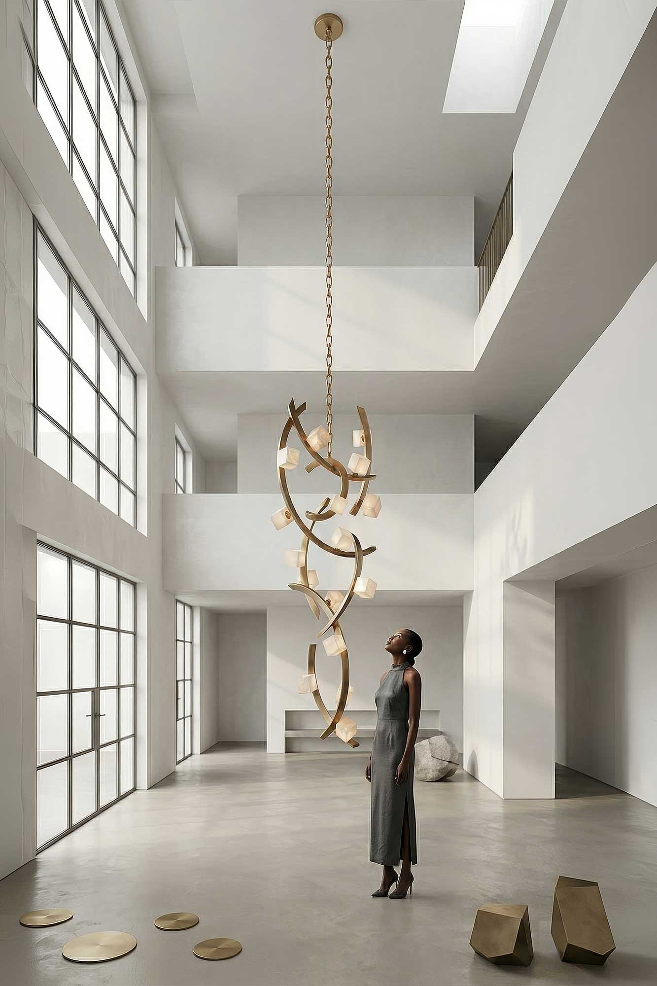A woman in a gray dress stands in a spacious, minimalist room with large windows, looking up at a modern gold chandelier suspended from the high ceiling. Gold geometric objects are on the floor nearby.