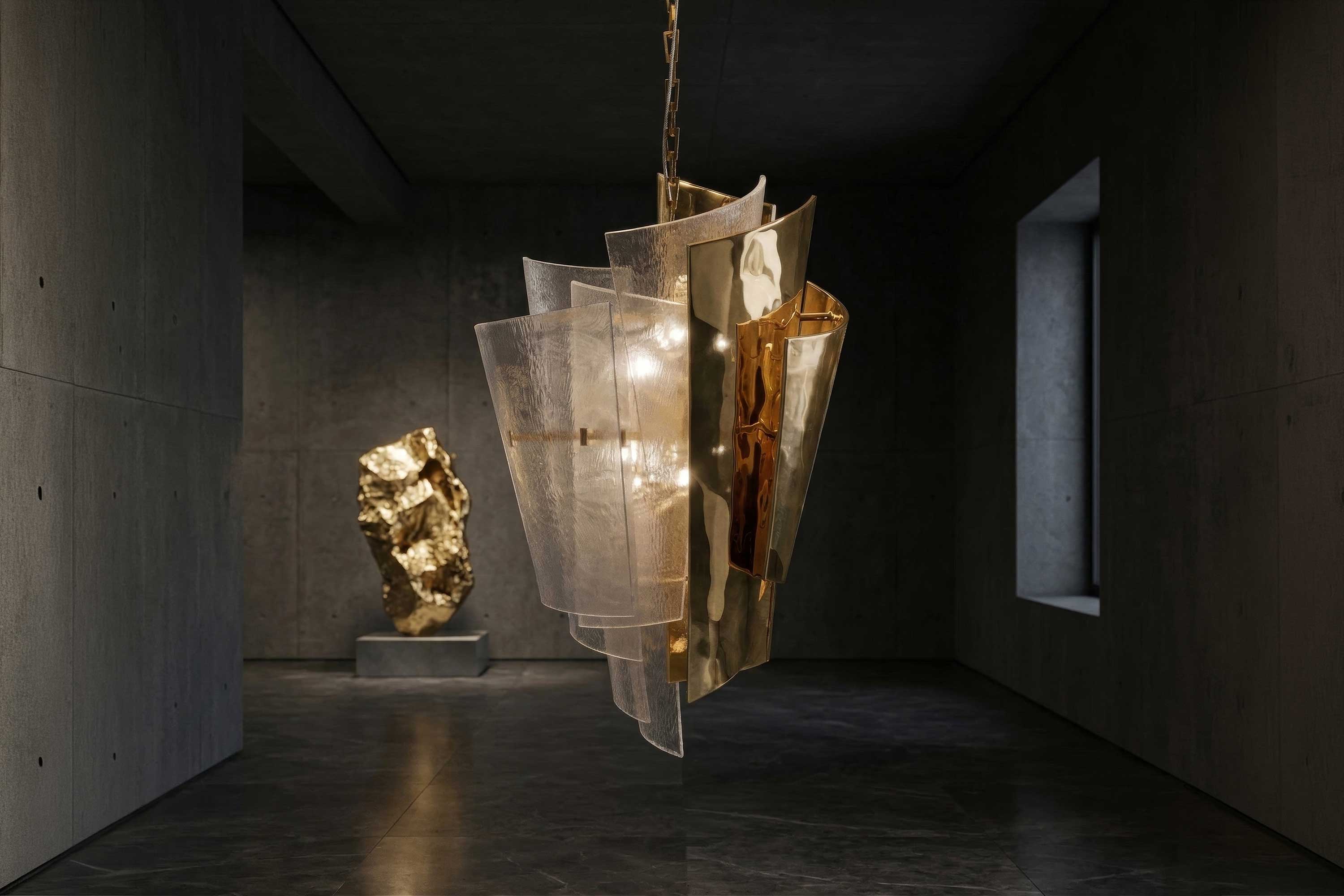 A modern pendant light with layered metal and glass panels hangs in a dim, minimalist room with concrete walls and floors. A gold abstract sculpture is displayed in the background on a pedestal.