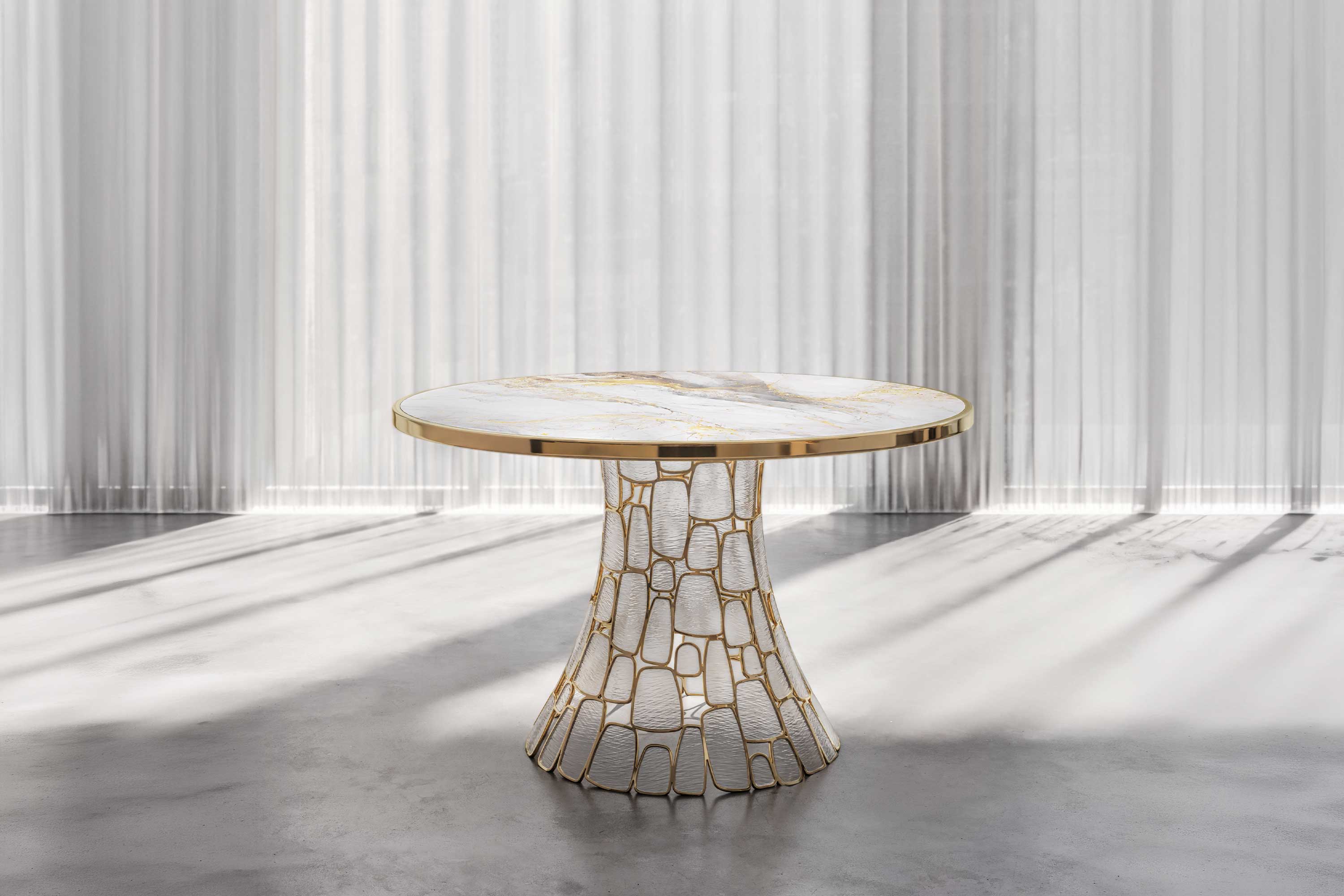 Round table with a white marble top and gold trim, supported by a sculptural gold and white base, standing on a polished concrete floor with sheer curtains and sunlight in the background.