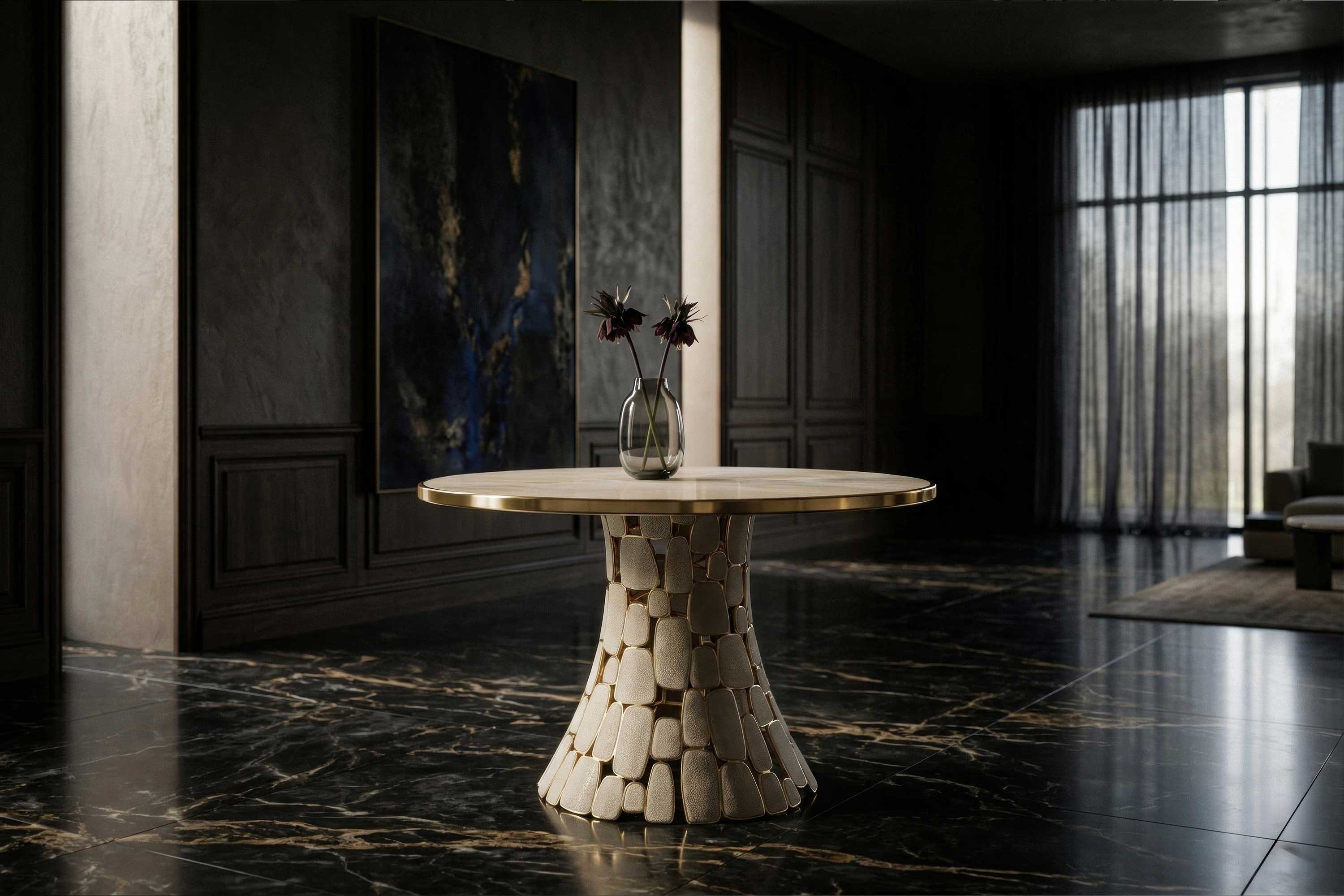 A round table with a stone-like base and a gold rimmed top sits on a dark marble floor in a modern room with dark walls, large windows, and sheer curtains. A glass vase with two dark flowers is on the table.