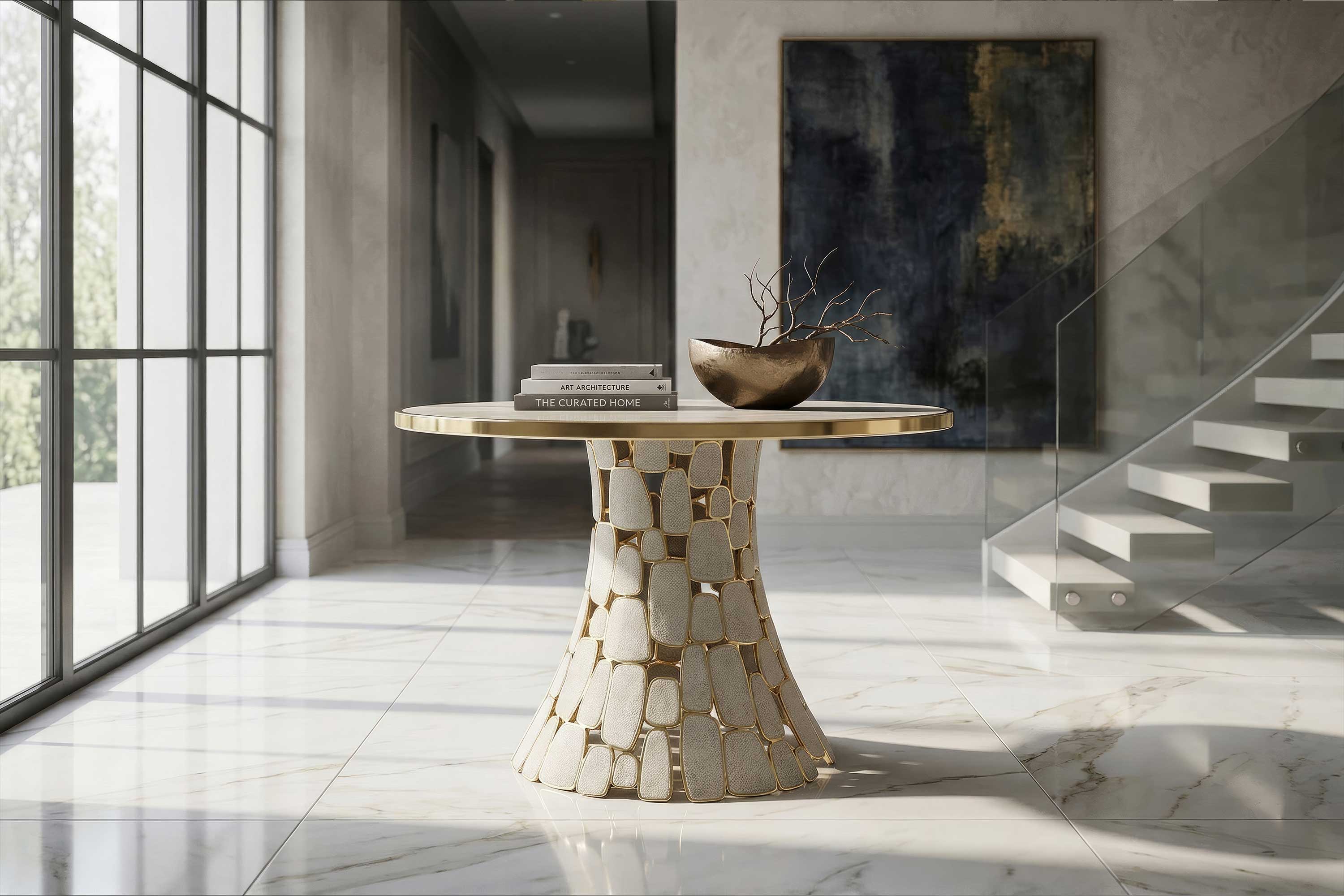 A round table with a stone base and gold trim holds books and a decorative bowl. The table is on a marble floor near large windows, with a staircase and abstract art in the background.