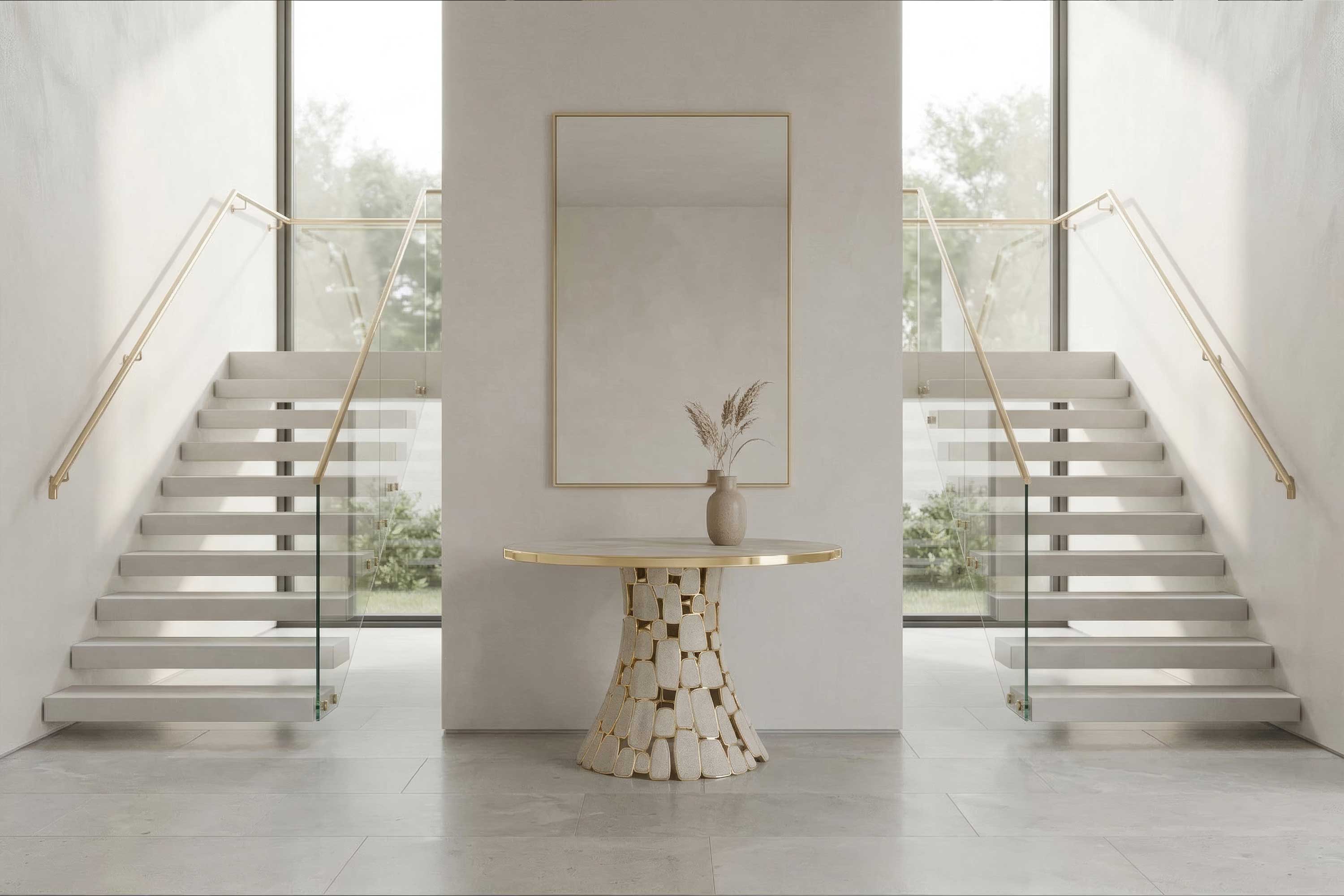 A modern entryway with a round table holding a beige vase and dried plants, a large rectangular mirror above, and two glass-railed staircases on either side, with large windows showing greenery outside.