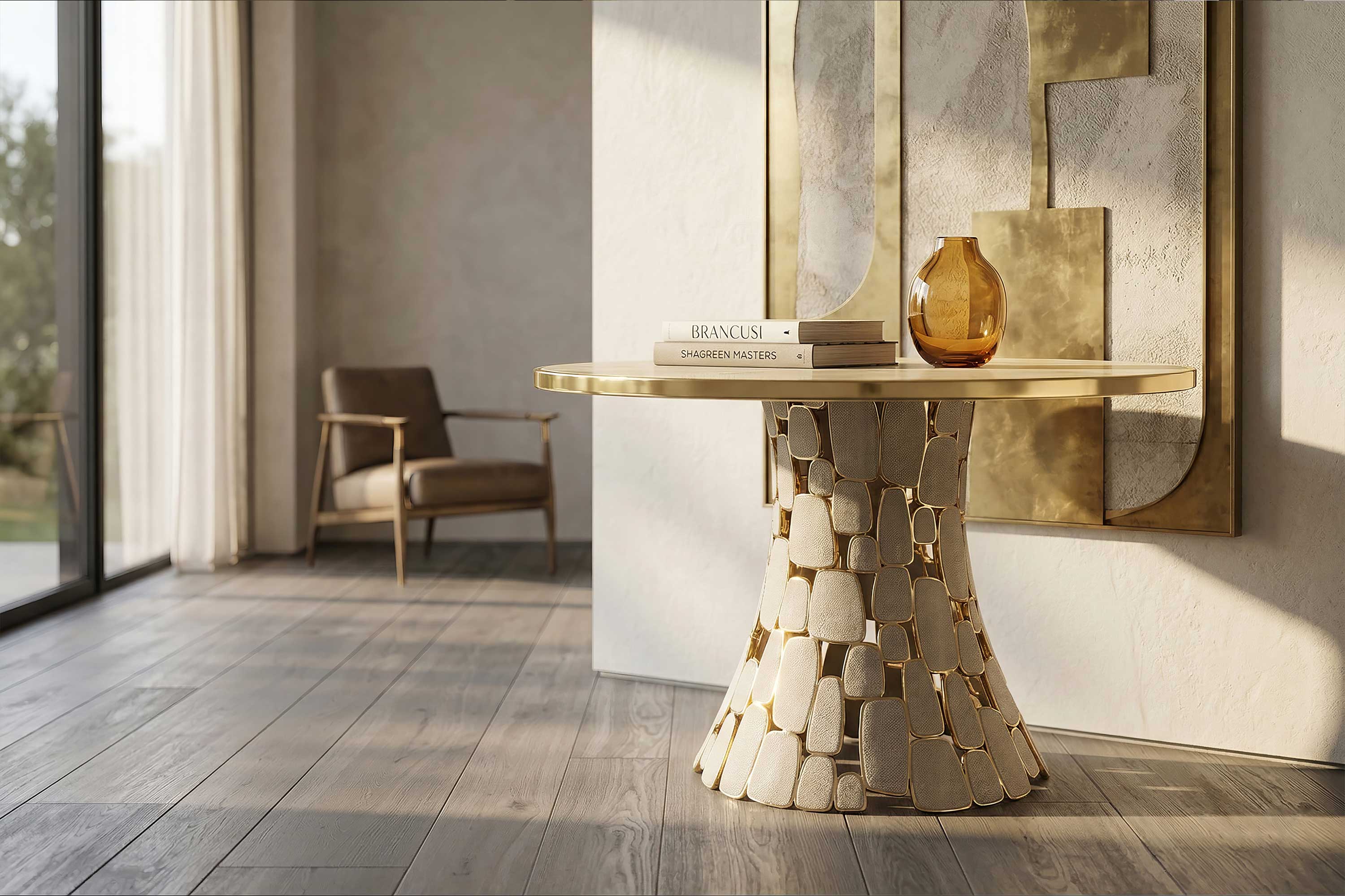 A round gold table with a unique block-patterned base stands on wooden flooring. On it are stacked books and an amber glass vase. Behind, theres a modern armchair and abstract gold wall art. Sunlight streams through large windows.