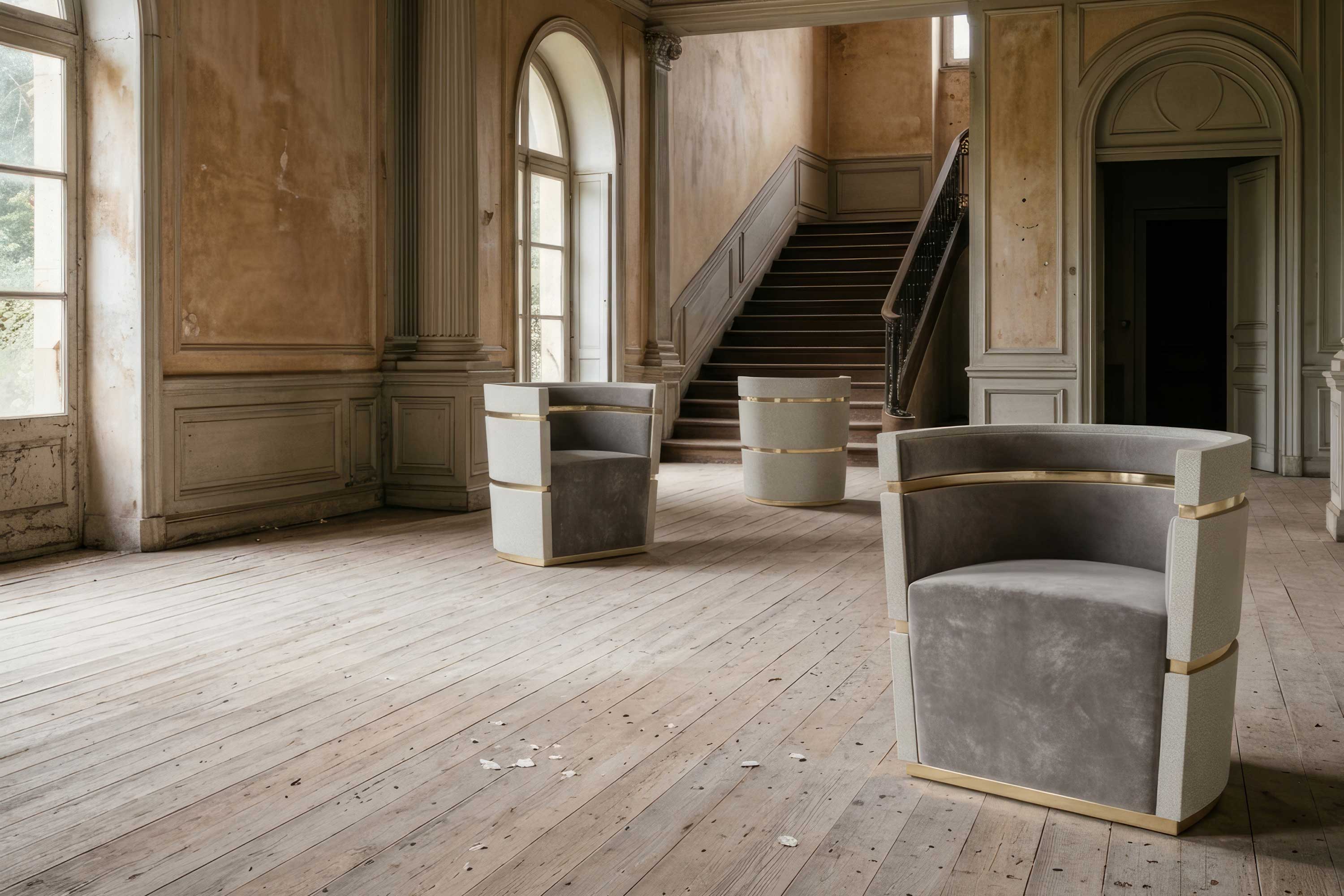 Three modern gray armchairs with gold accents are placed on a worn wooden floor in a large, empty room with peeling beige walls, tall windows, and a wooden staircase in the background.