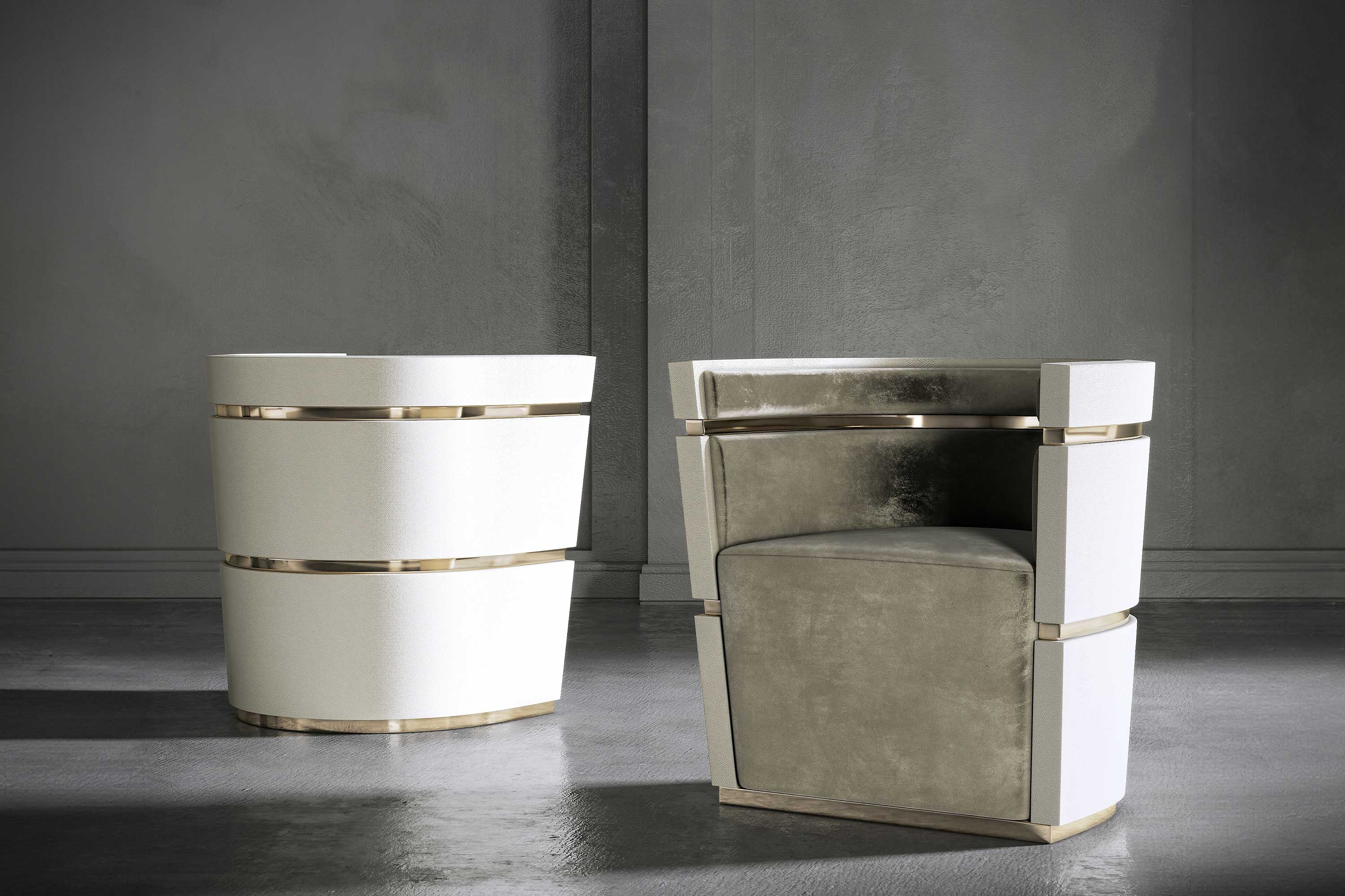 Two modern armchairs with segmented, geometric designs. One chair is upholstered in cream fabric, and the other in a light grayish velvet, both featuring metallic trim accents and set against a concrete wall and floor.