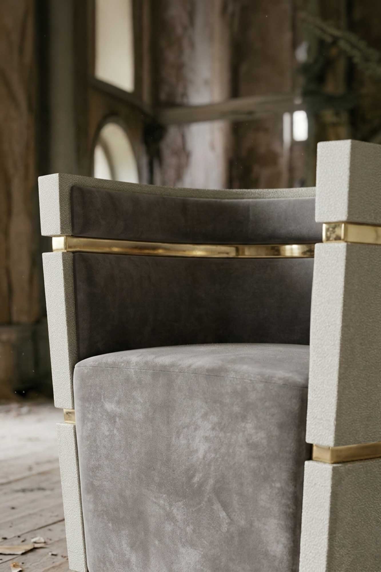 Modern accent chair with grey upholstery and gold metal accents, positioned in a rustic room with weathered walls, arched windows, and wooden floors. Only part of the chair is visible in the foreground.