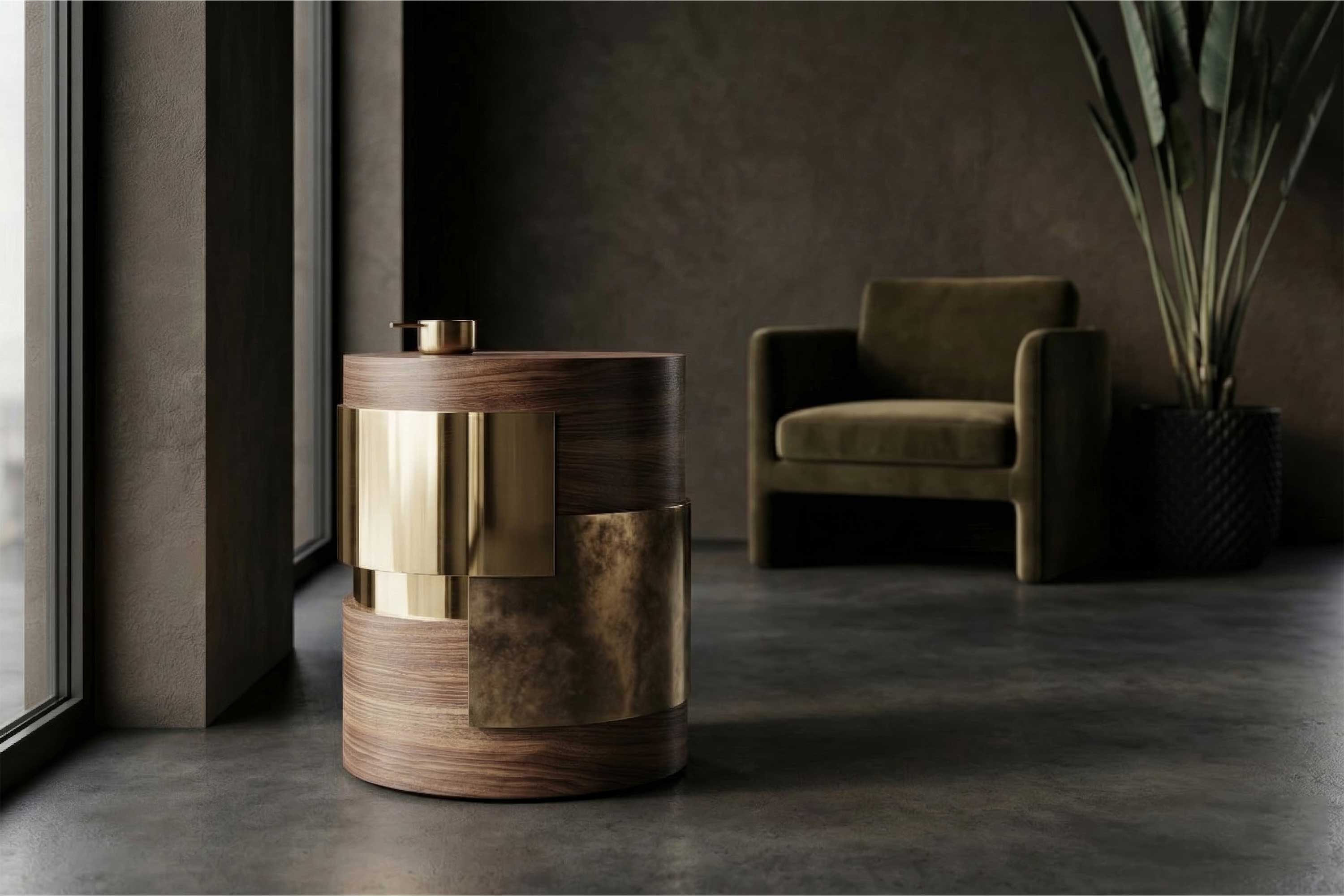 A round, modern side table with wood and bronze accents sits near a large window. An olive green armchair and tall plant are in the background, set against dark walls and a concrete floor.