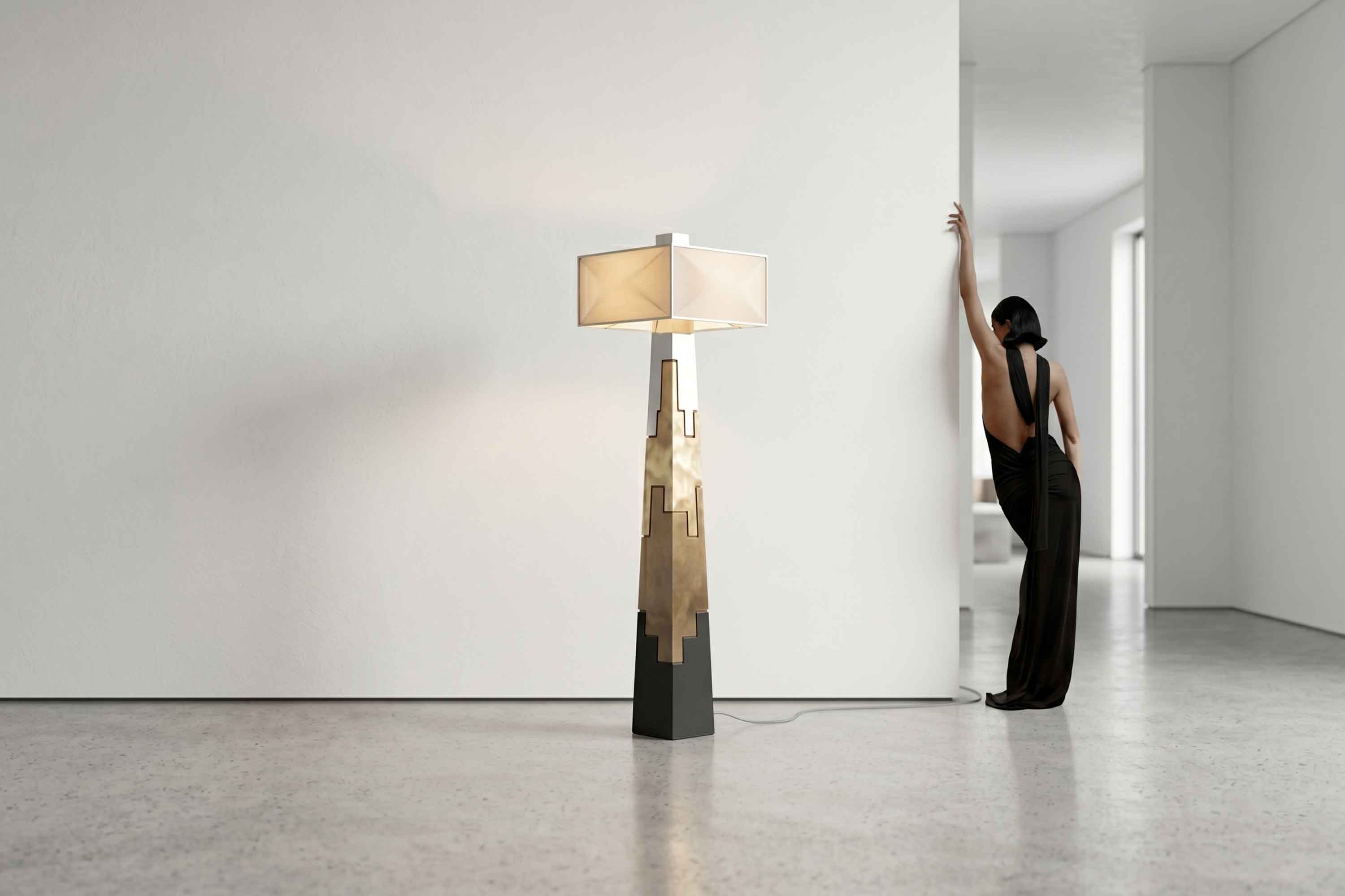 A modern floor lamp with a geometric base and rectangular shade stands in a minimalist room, channeling an Artdecoo aesthetic. A person in a black dress leans against a wall in the background, partially turned away. The light-colored, polished floor enhances the clean atmosphere.