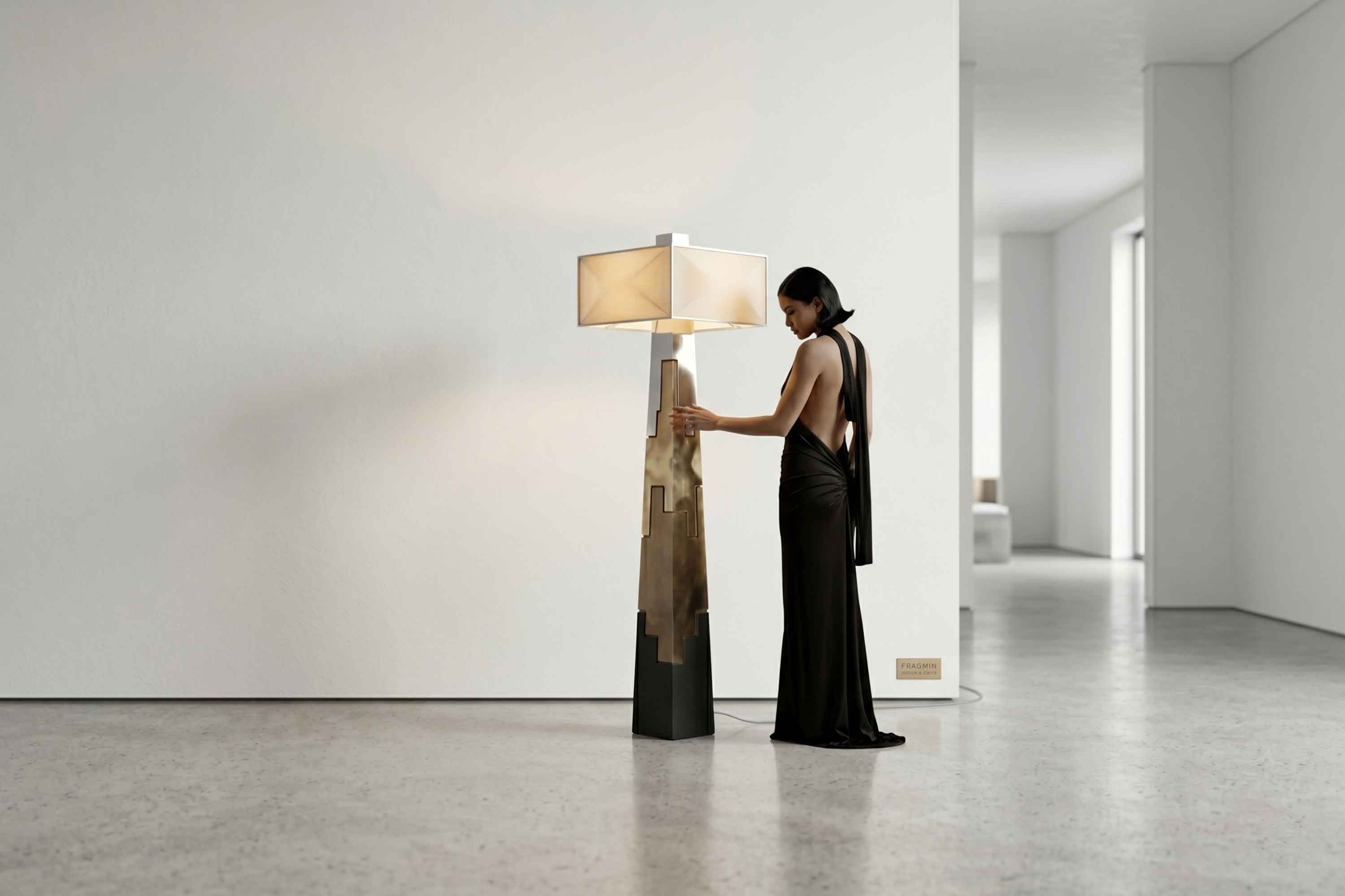 A woman in a black dress stands next to a modern, handmade floor lamp with a geometric base and a square lampshade in a minimalist, spacious room with white walls and an open doorway in the background.