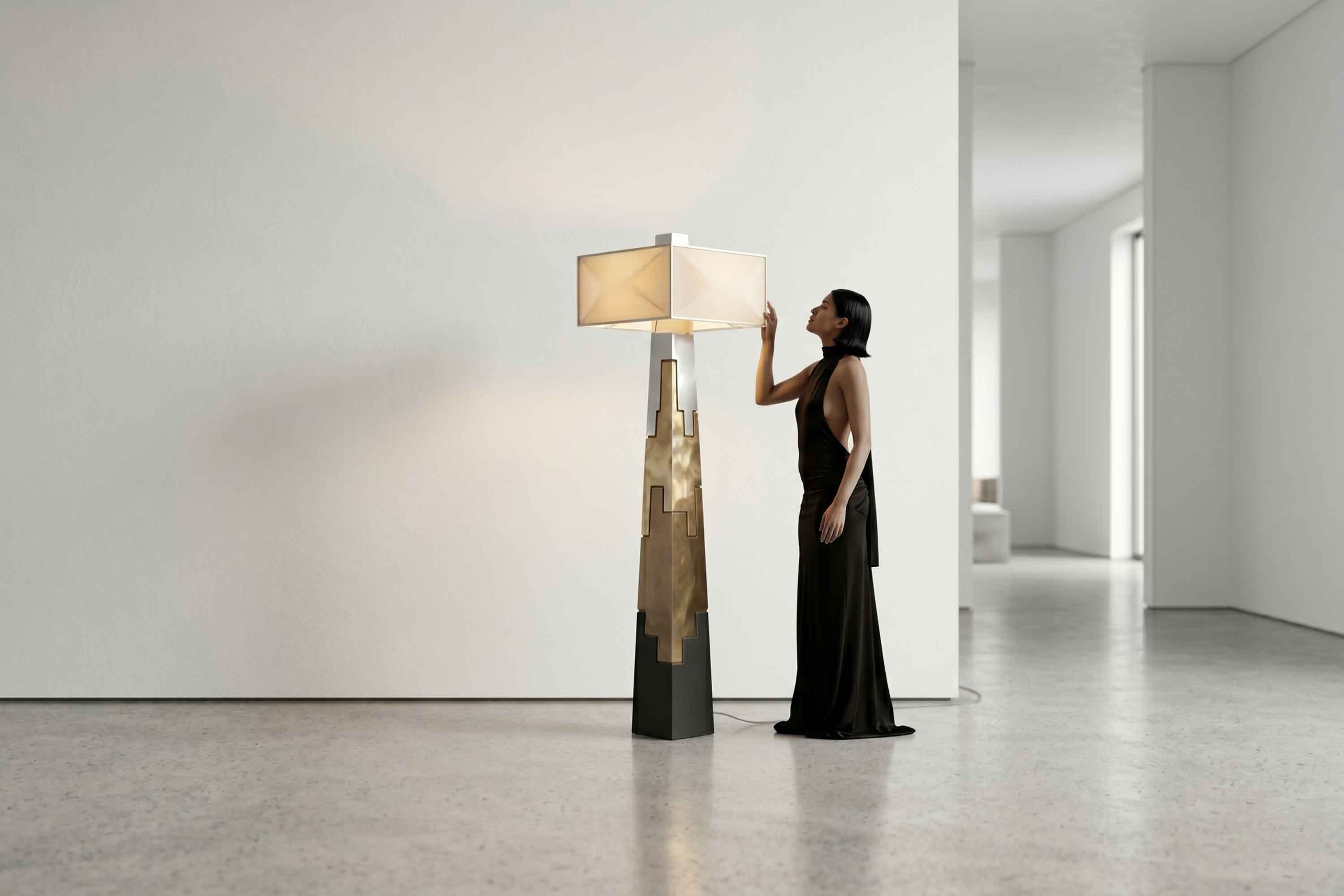 A woman in a long black dress stands in a minimalist room, gently touching a modern floor lamp with a geometric Bronze and black base and a white lampshade. The room features white walls and a polished concrete floor, complemented by touches of Bespoke Furniture.