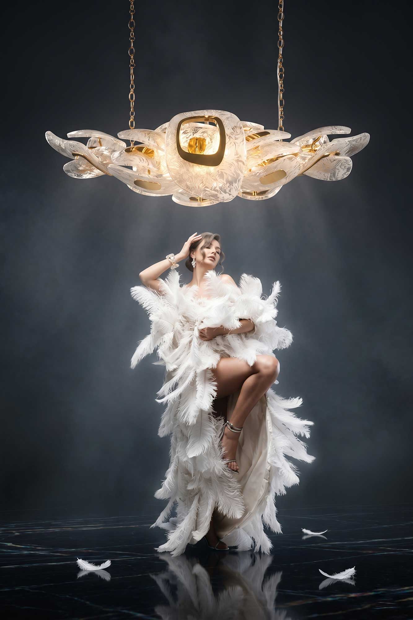 A woman in a dramatic white feathered gown poses under a large, ornate light fixture with a gold center and sculpted translucent wings. The background is dark, and a few white feathers lie on the shiny floor.