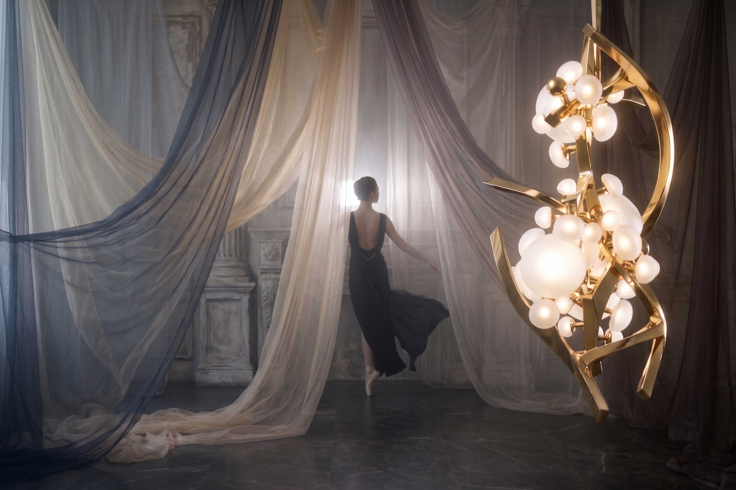 A person in a long, dark dress stands near sheer curtains in a dim room with marble walls. A modern gold and white chandelier hangs in the foreground. Light filters softly from behind the figure.