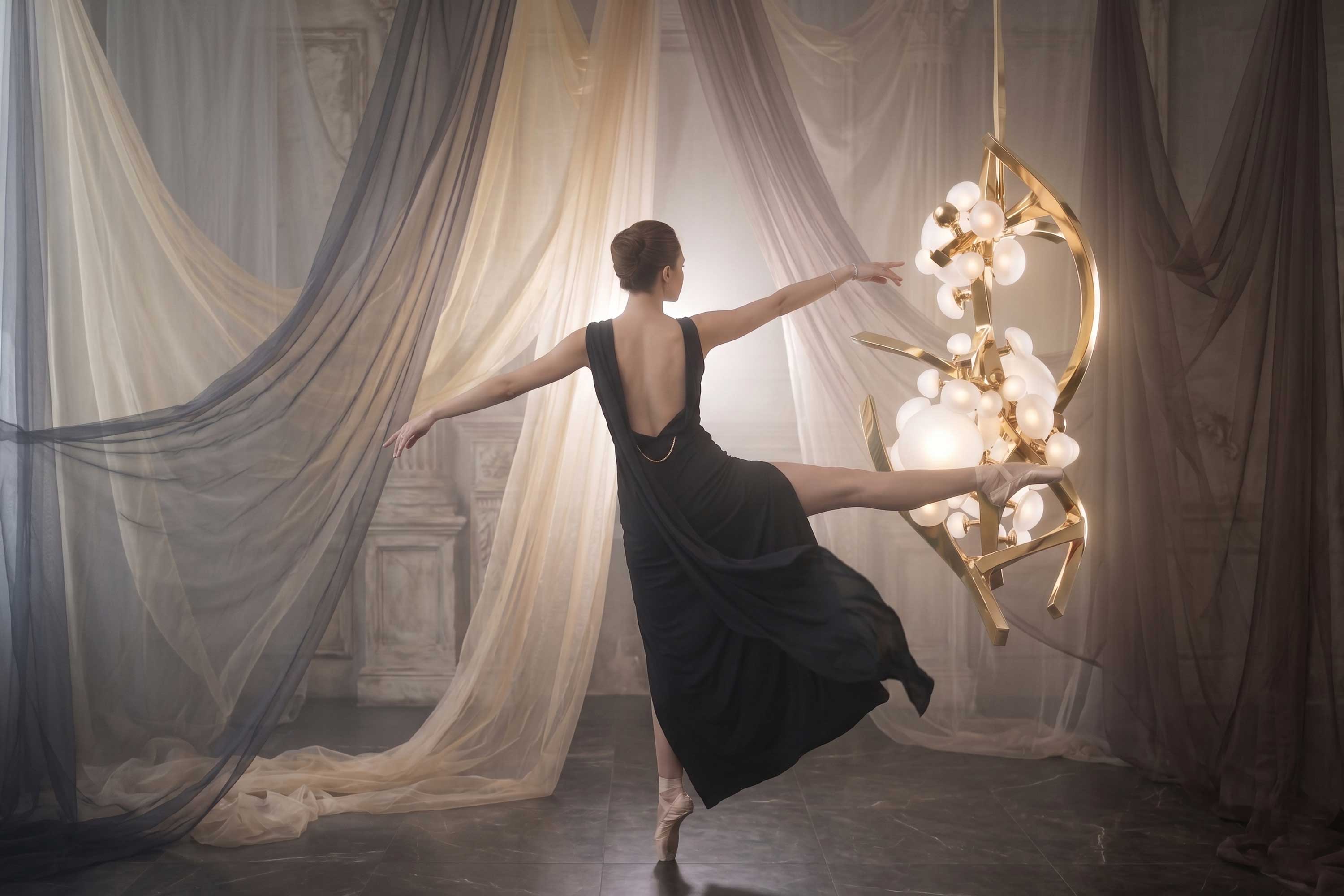 A ballerina in a black dress balances on one foot, extending her leg to touch a modern chandelier. She is surrounded by soft, sheer drapes in a dimly lit room with marble flooring.