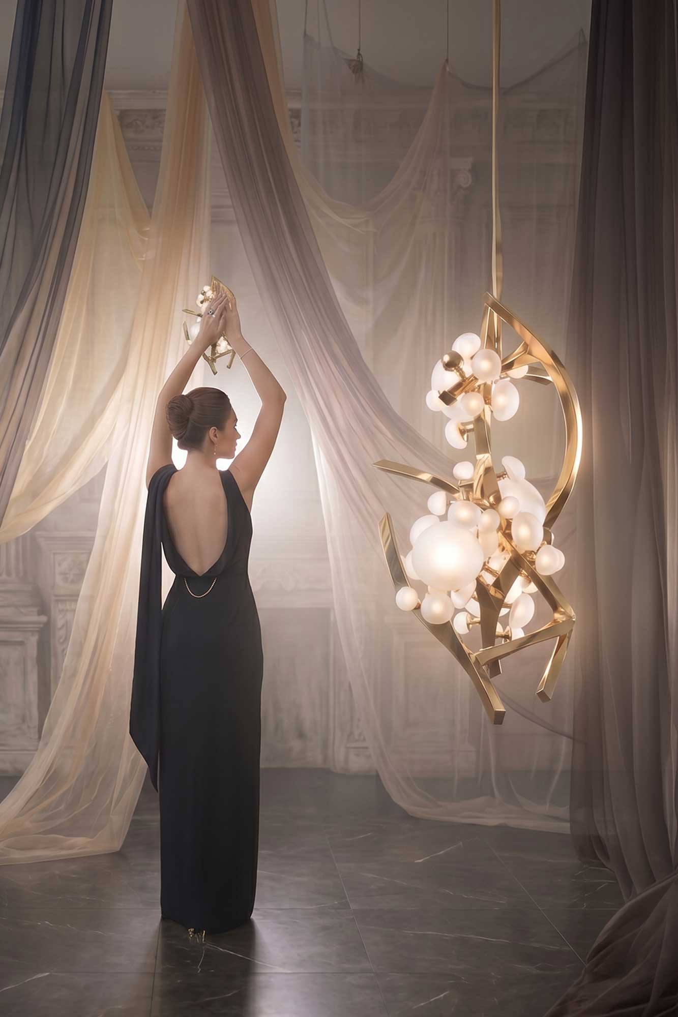 A woman in a backless black gown stands with arms raised, facing sheer drapes and ornate walls. Beside her, a modern chandelier with gold branches and glowing orbs hangs, creating a soft, elegant atmosphere.