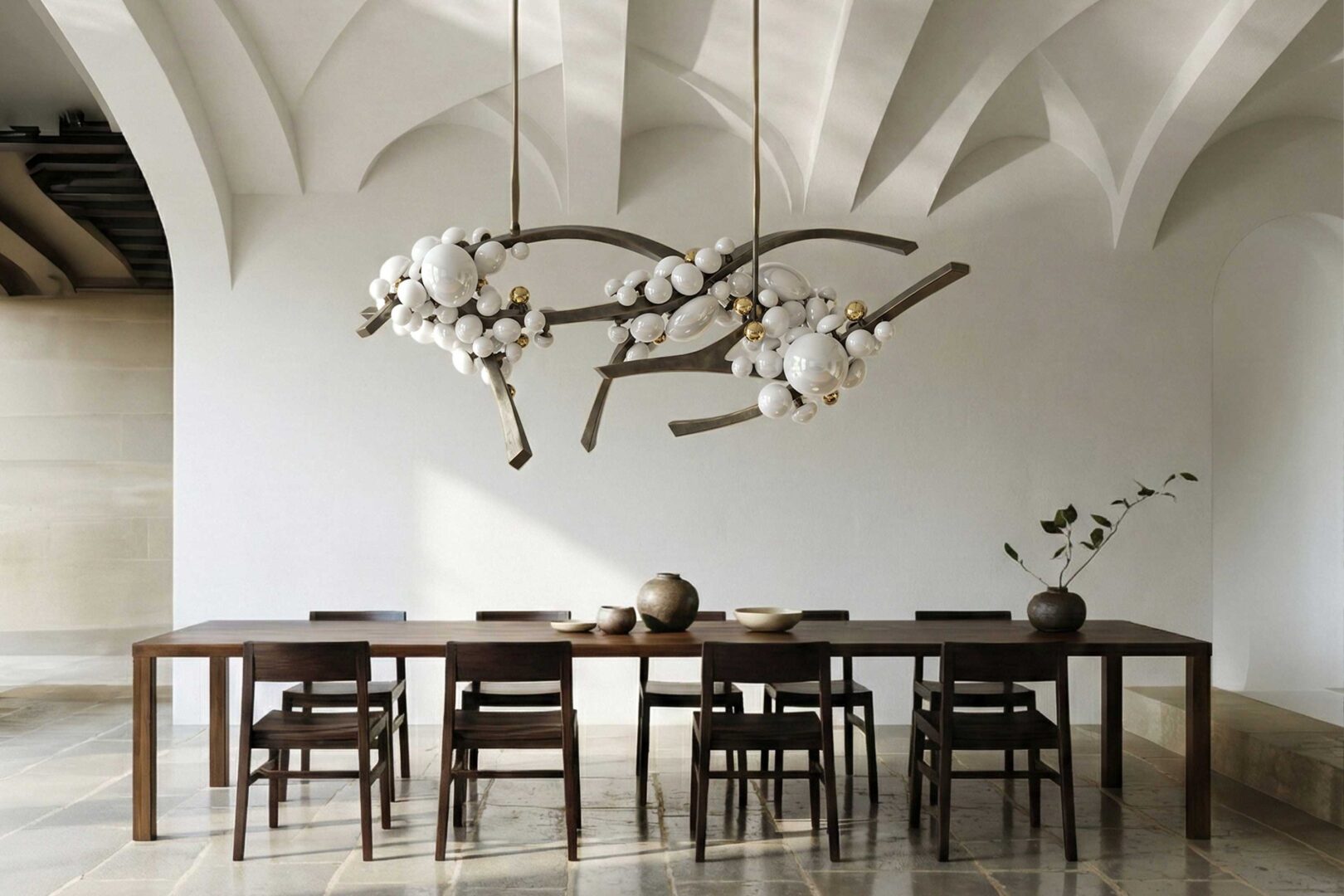 A long wooden dining table with eight chairs is set beneath two modern chandeliers. The room has vaulted white ceilings, light stone floors, and minimalist decor, including a few ceramic vases and a branch in a vase.