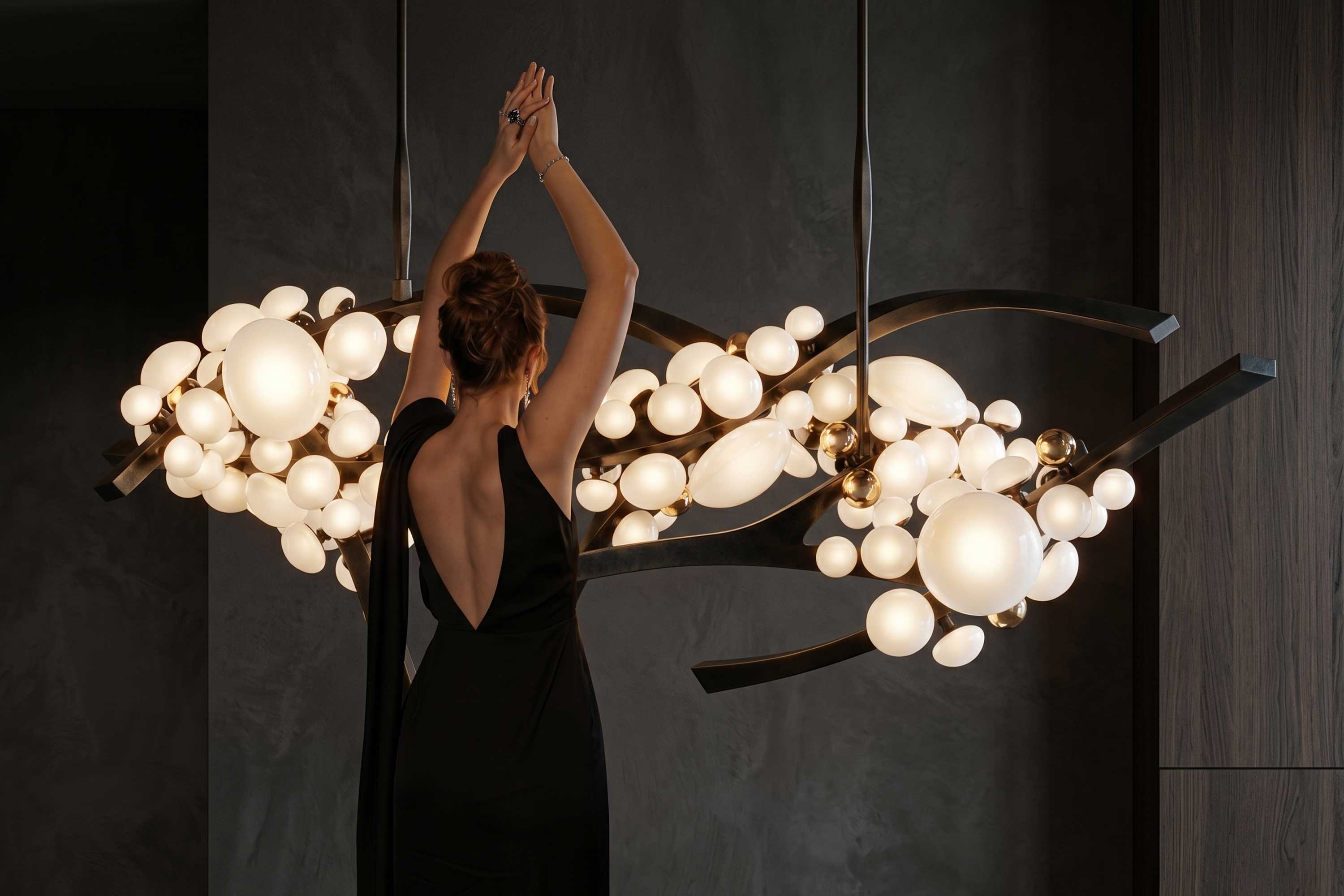 A woman in a black dress stands with her arms raised near a modern chandelier featuring spherical lights and dark metal branches, set against a dark background.