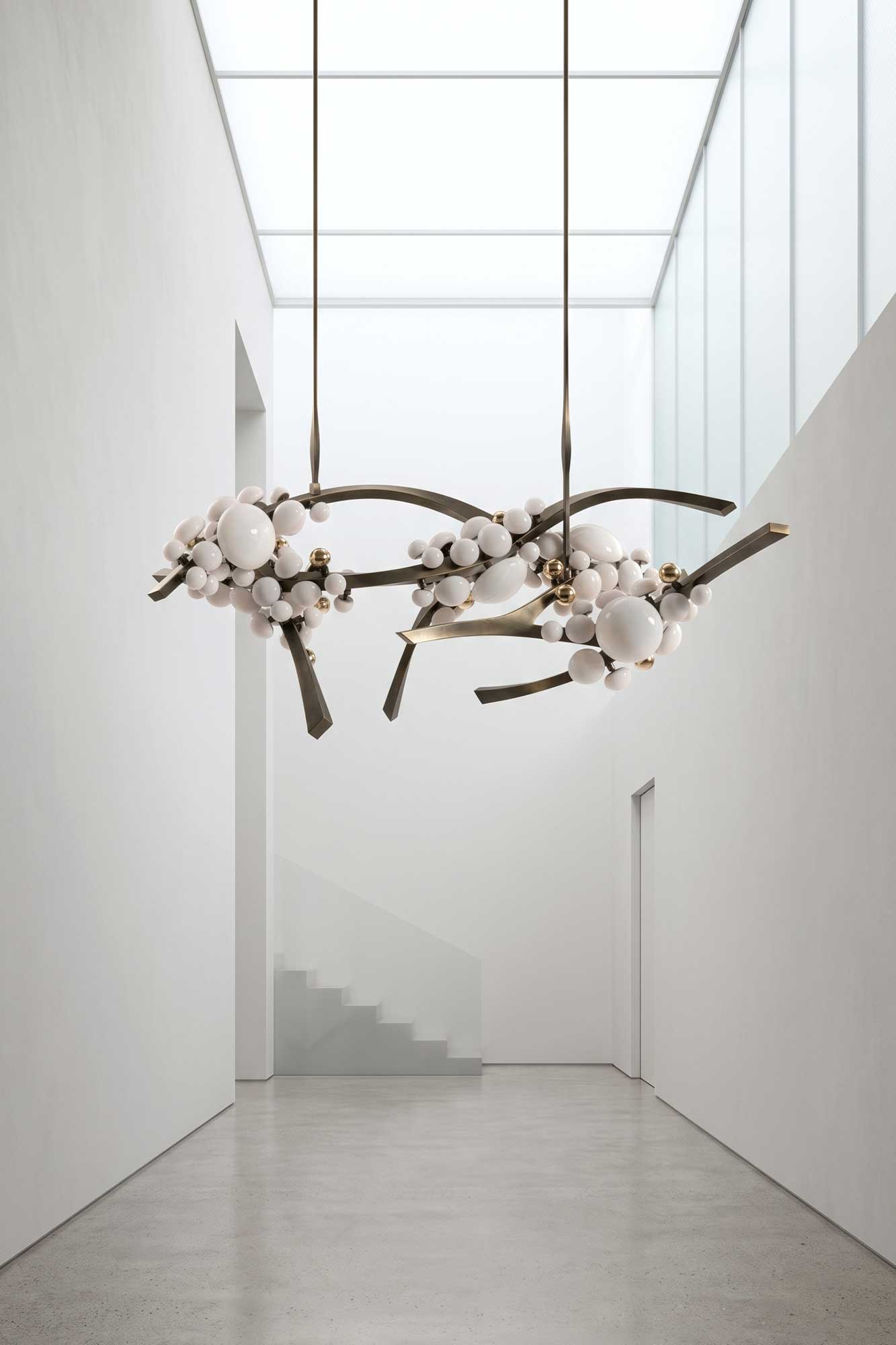 A minimalist white interior with a staircase, glass ceiling, and two modern chandeliers featuring white and bronze spherical elements and branching metal rods.