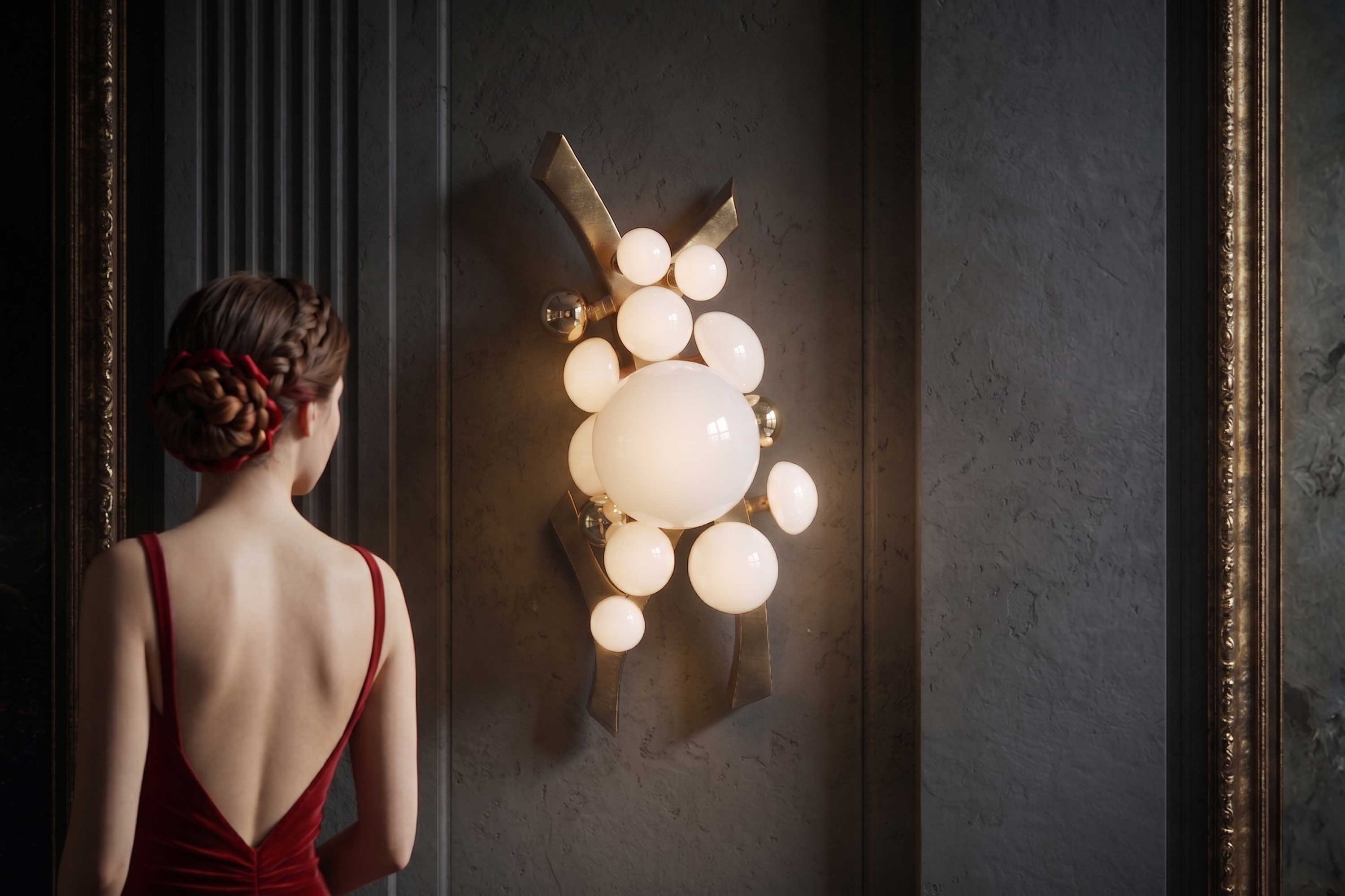 A woman in a red dress with an open back stands facing a gray wall adorned with a modern light fixture made of spherical white bulbs, bronze accents, and gold elements. The setting appears elegant and softly lit.