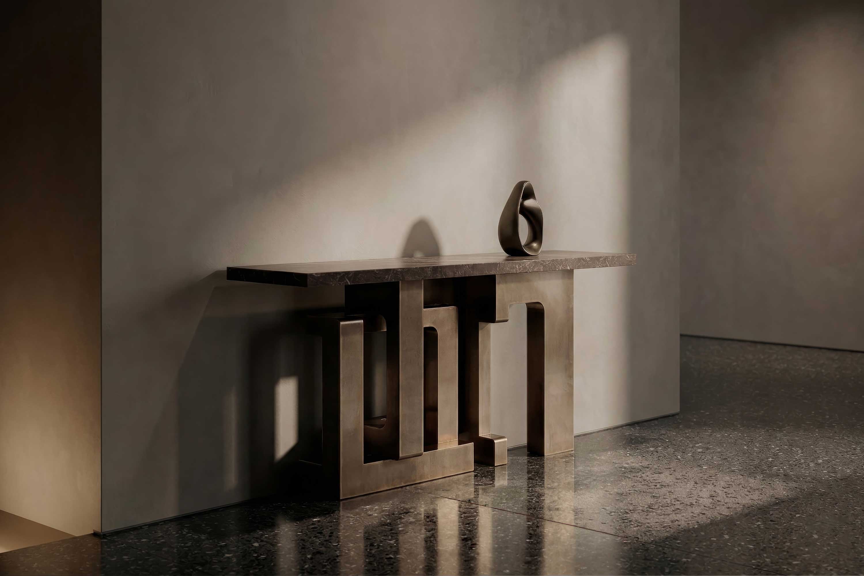 A modern console table with a geometric bronze base stands against a beige wall. A single abstract vase is placed on top. Light casts soft shadows onto the floor and wall, highlighting the tables design.