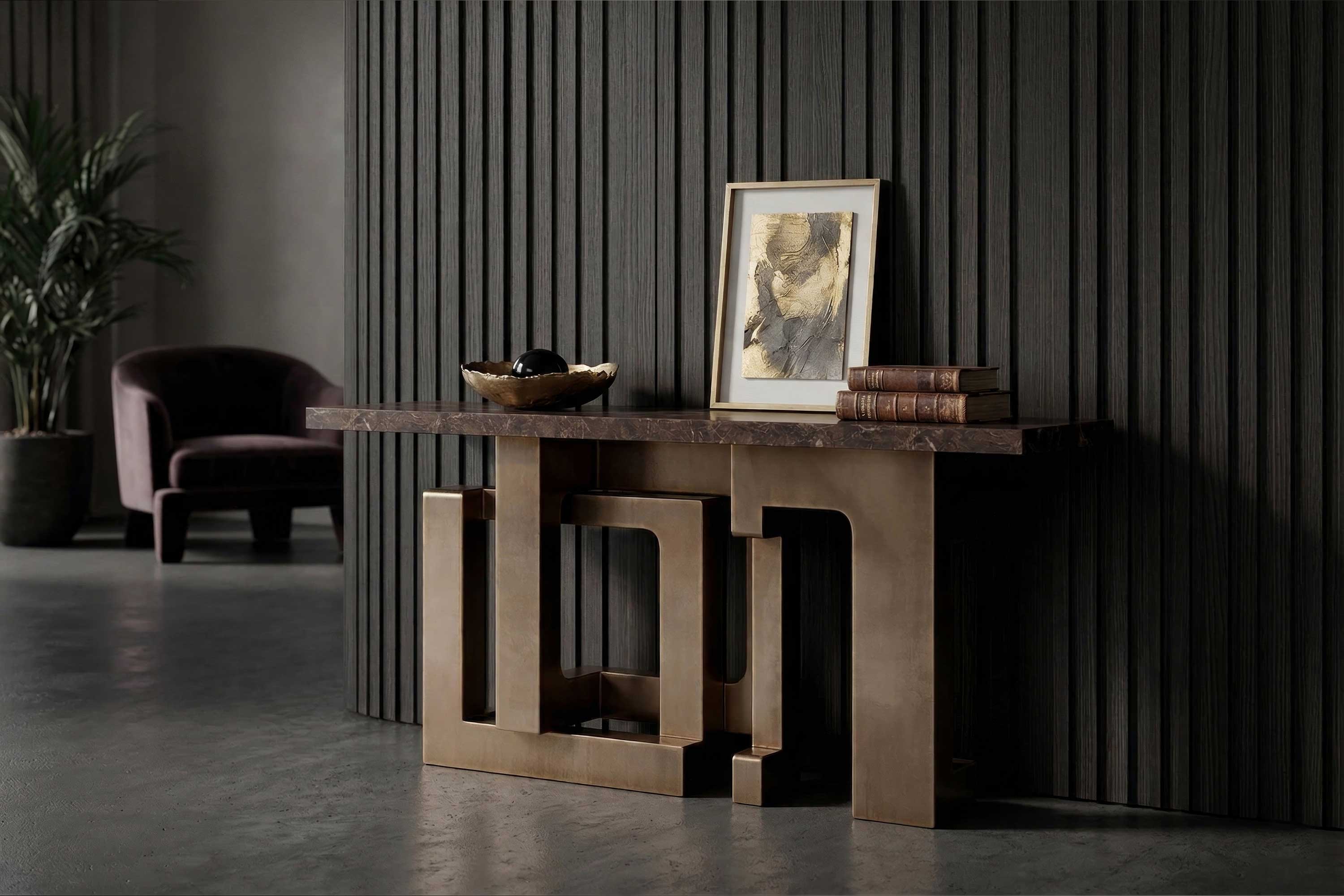A modern console table with a geometric gold base and marble top holds a bowl, a framed abstract artwork, and two stacked books. A dark armchair and a potted plant are in the background against a ribbed wall.