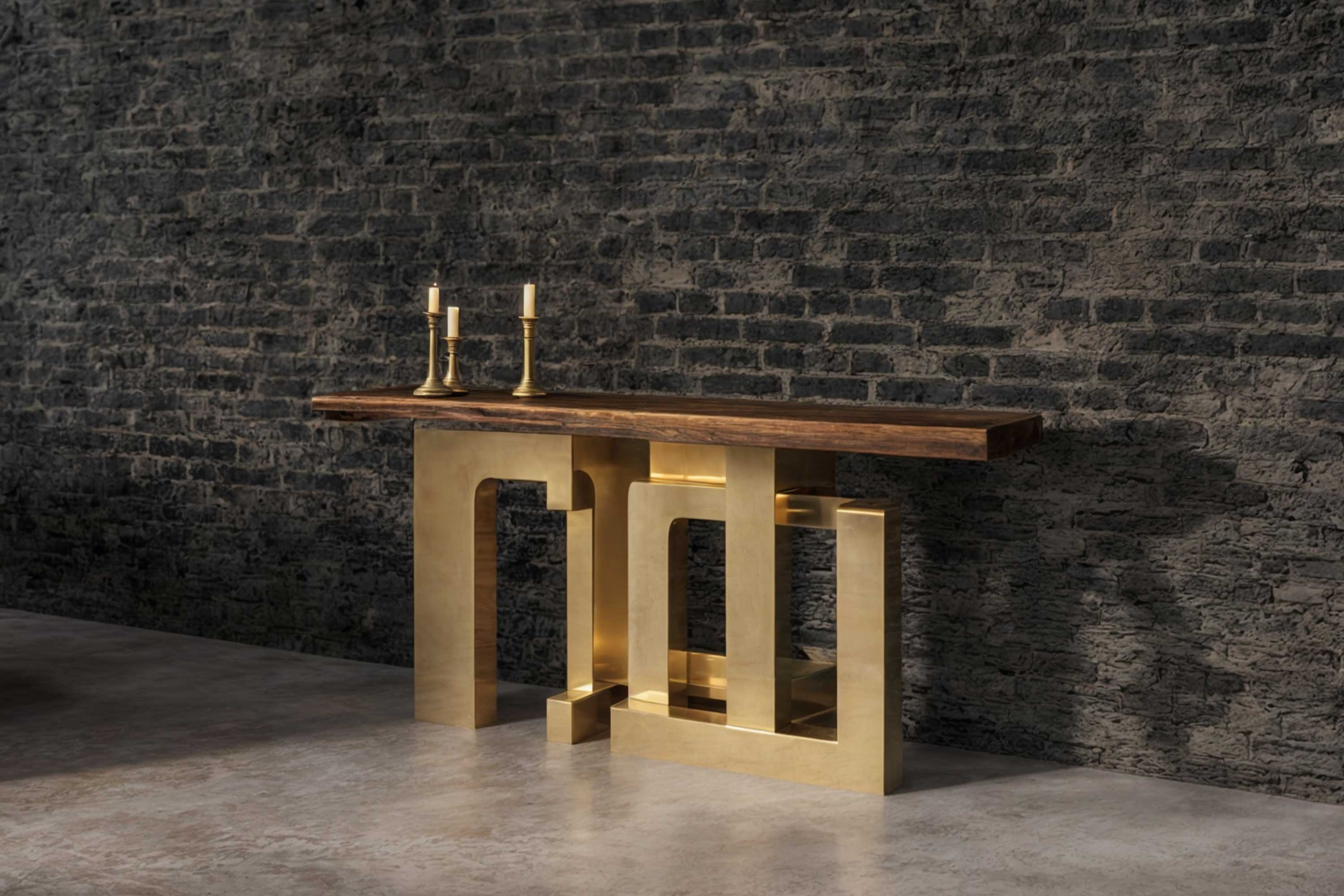 A modern console table with a wooden top and geometric gold metal base is placed against a textured dark brick wall. Two gold candlesticks sit on the left side of the table. The floor is a smooth concrete surface.
