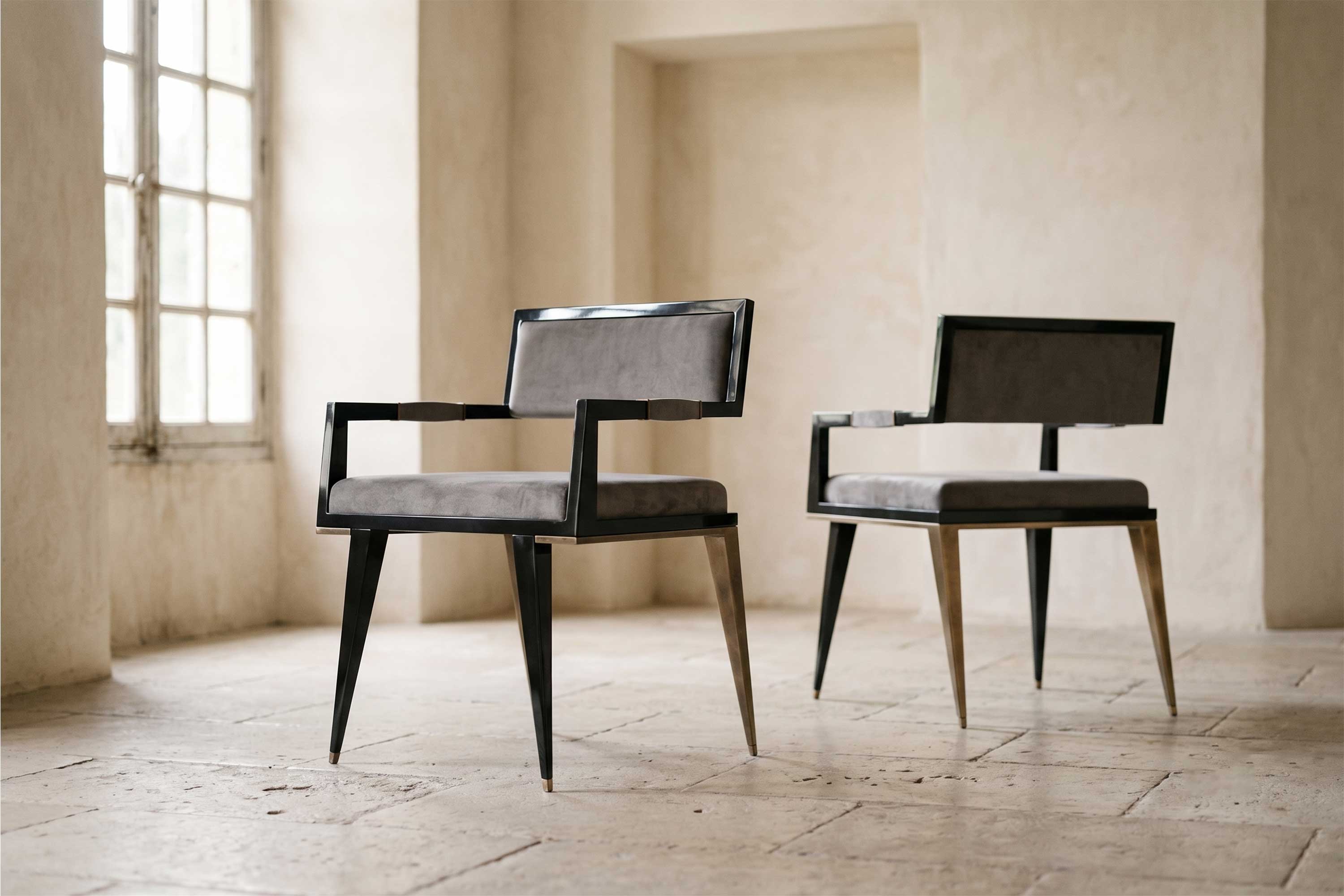 Two modern armchairs with black frames and grey upholstered seats and backs sit on a stone tile floor in a neutral-toned room with large windows and natural light.