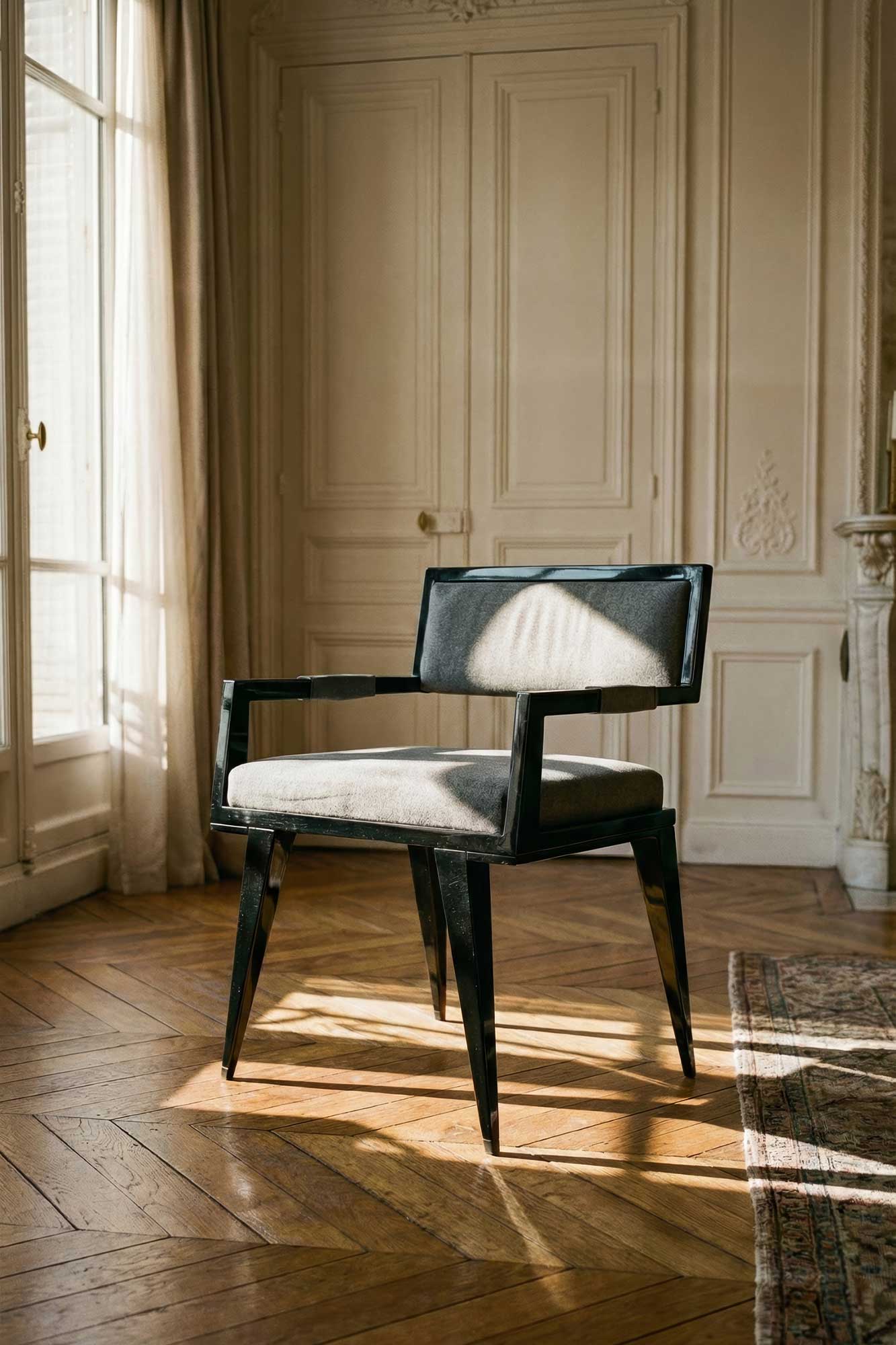 A modern black-framed chair with gray upholstery sits on a wooden herringbone floor in a sunlit room with cream-colored paneled walls, large windows, and a patterned rug partially visible.