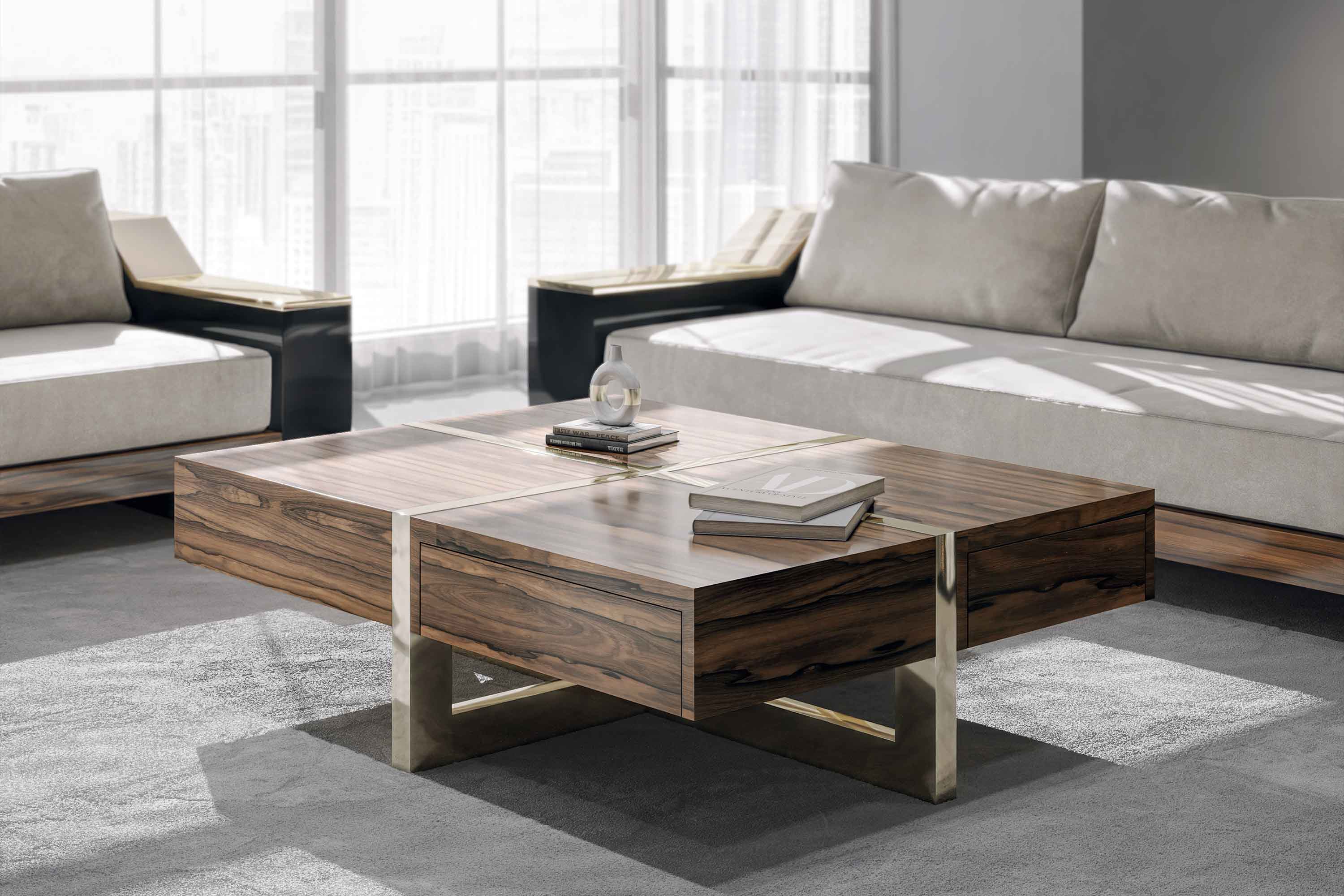 A modern living room with a large wooden coffee table, a few books, and a decorative object on top. Two light-colored sofas are positioned around the table. Large windows in the background let in natural light.