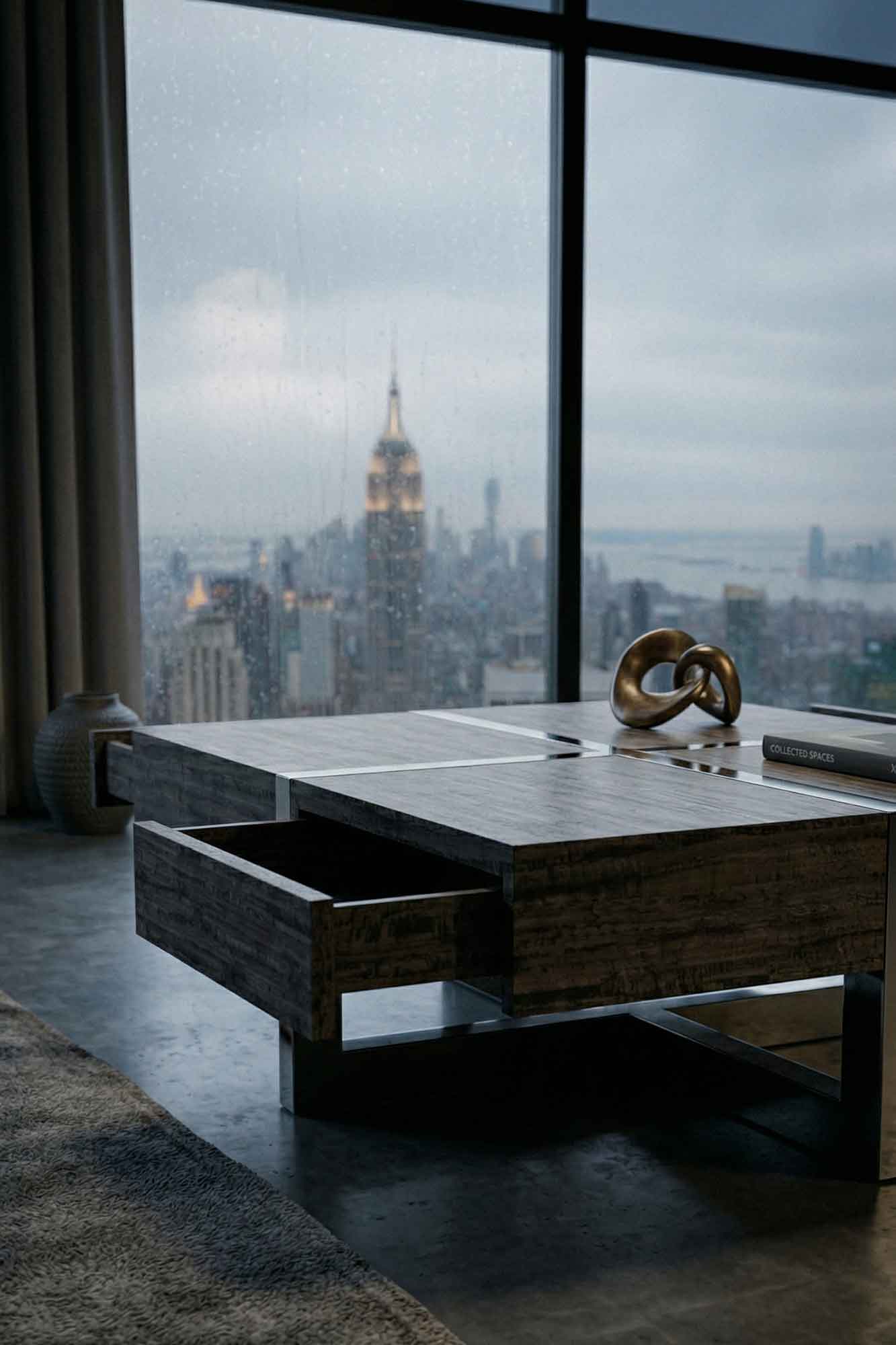 A modern coffee table with open drawers stands in front of a large window overlooking a rainy cityscape with the Empire State Building in the distance. A decorative object and book rest on the table.