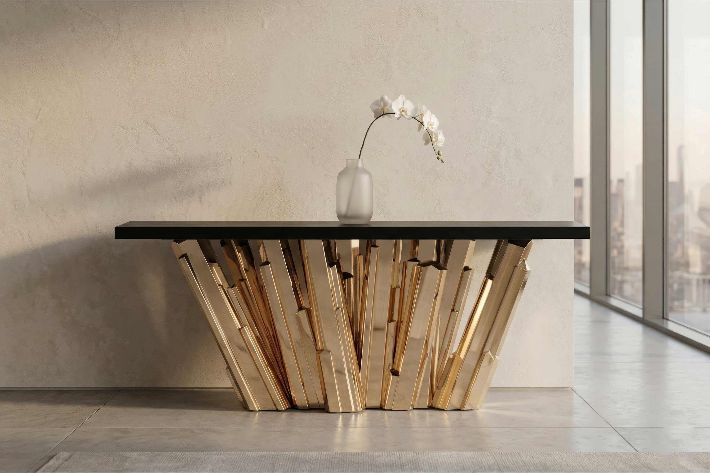 A sculptural table with gold, angular supports and a black top stands against a beige wall. A single white orchid in a frosted vase sits on the table. Large windows show a cityscape in the background.