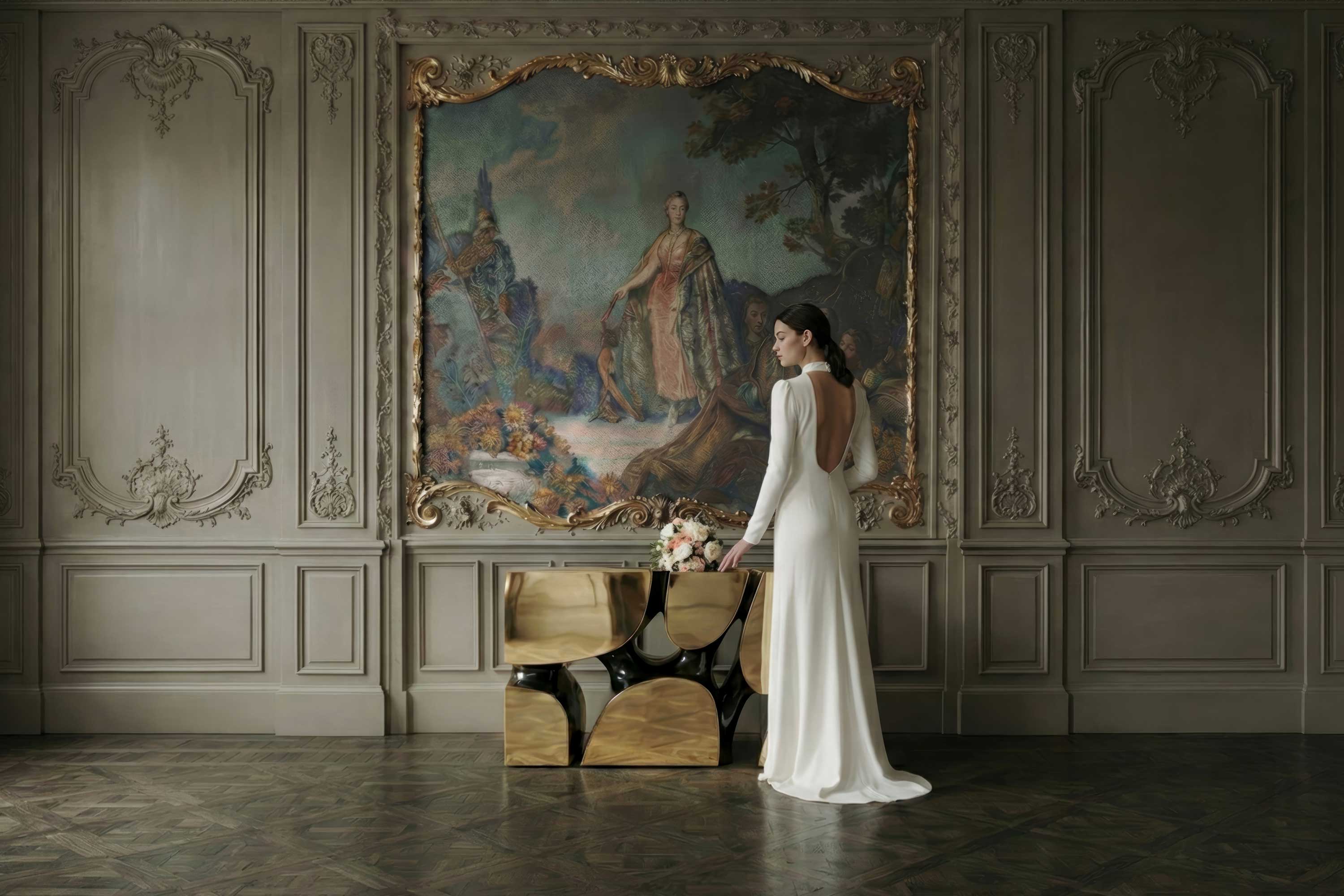 A woman in a long white dress stands beside a modern gold and black console table, holding a bouquet, in a formal room with ornate wall panels and a large classical painting.