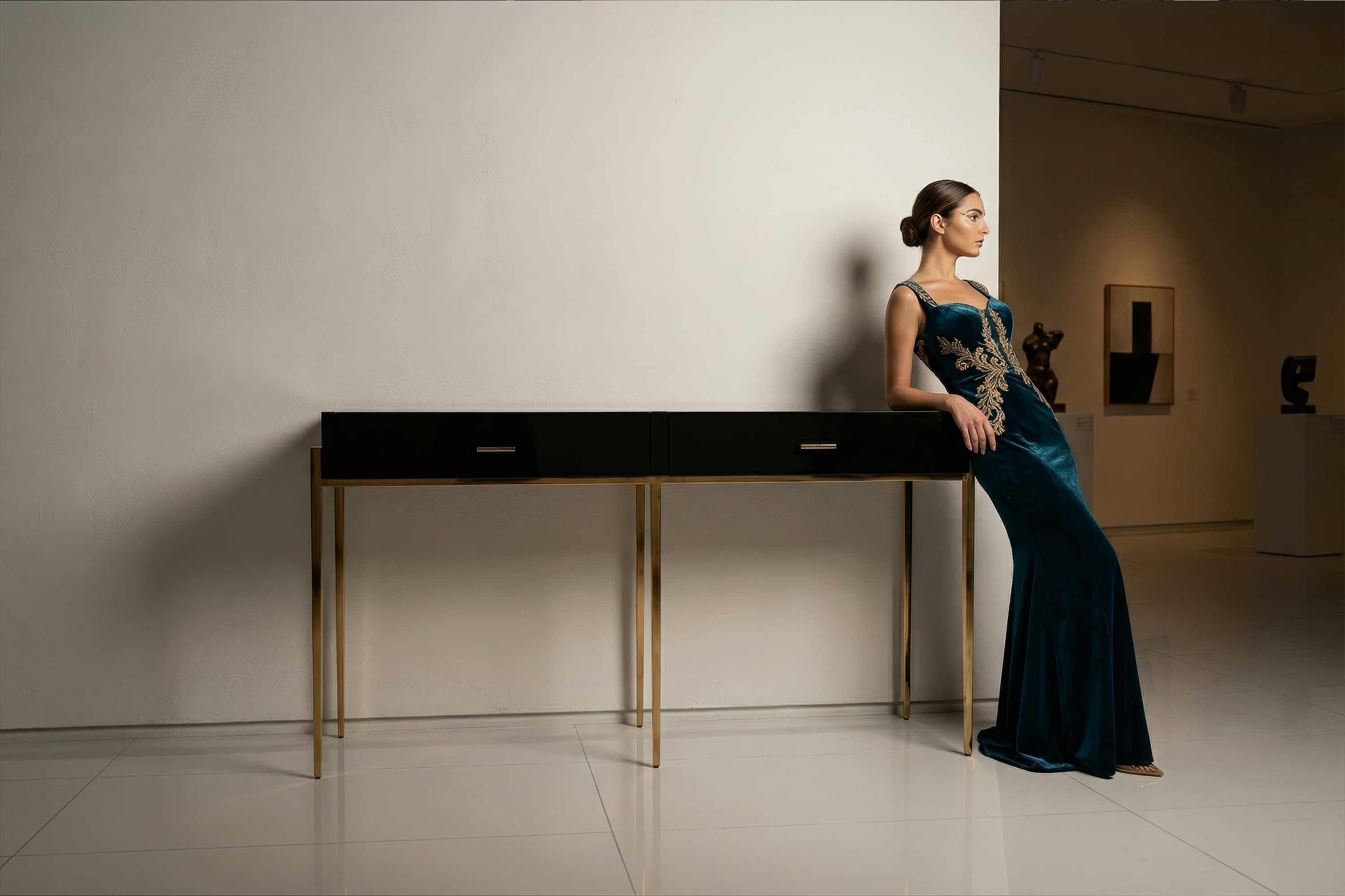 A woman in a long teal evening gown with gold embroidery leans against a black console table in a modern, minimalistic art gallery with white walls and glossy white floors.