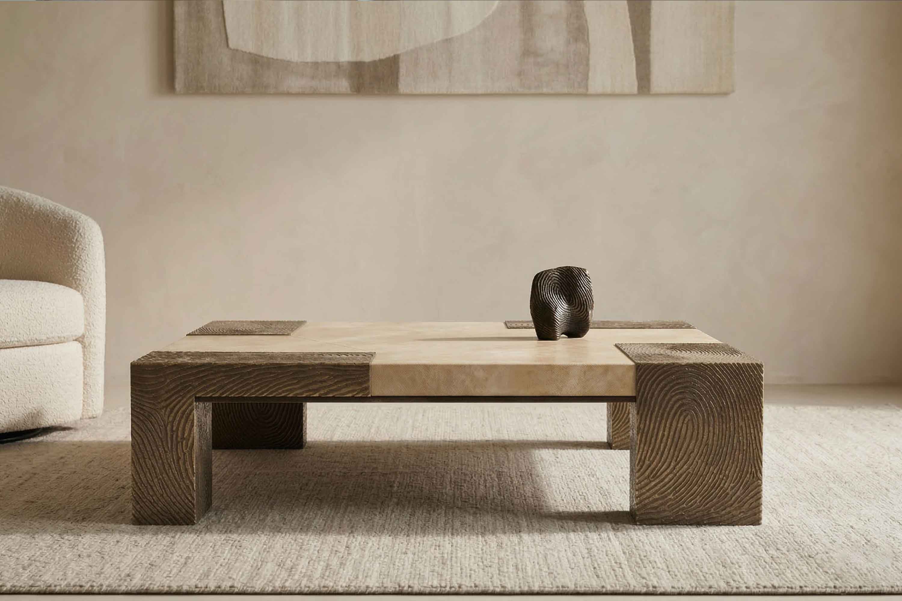 A low, rectangular coffee table with textured, dark wood legs and a smooth, light-colored top sits on a beige rug; a small, dark decorative object is placed on the table. Abstract artwork hangs on the wall behind.